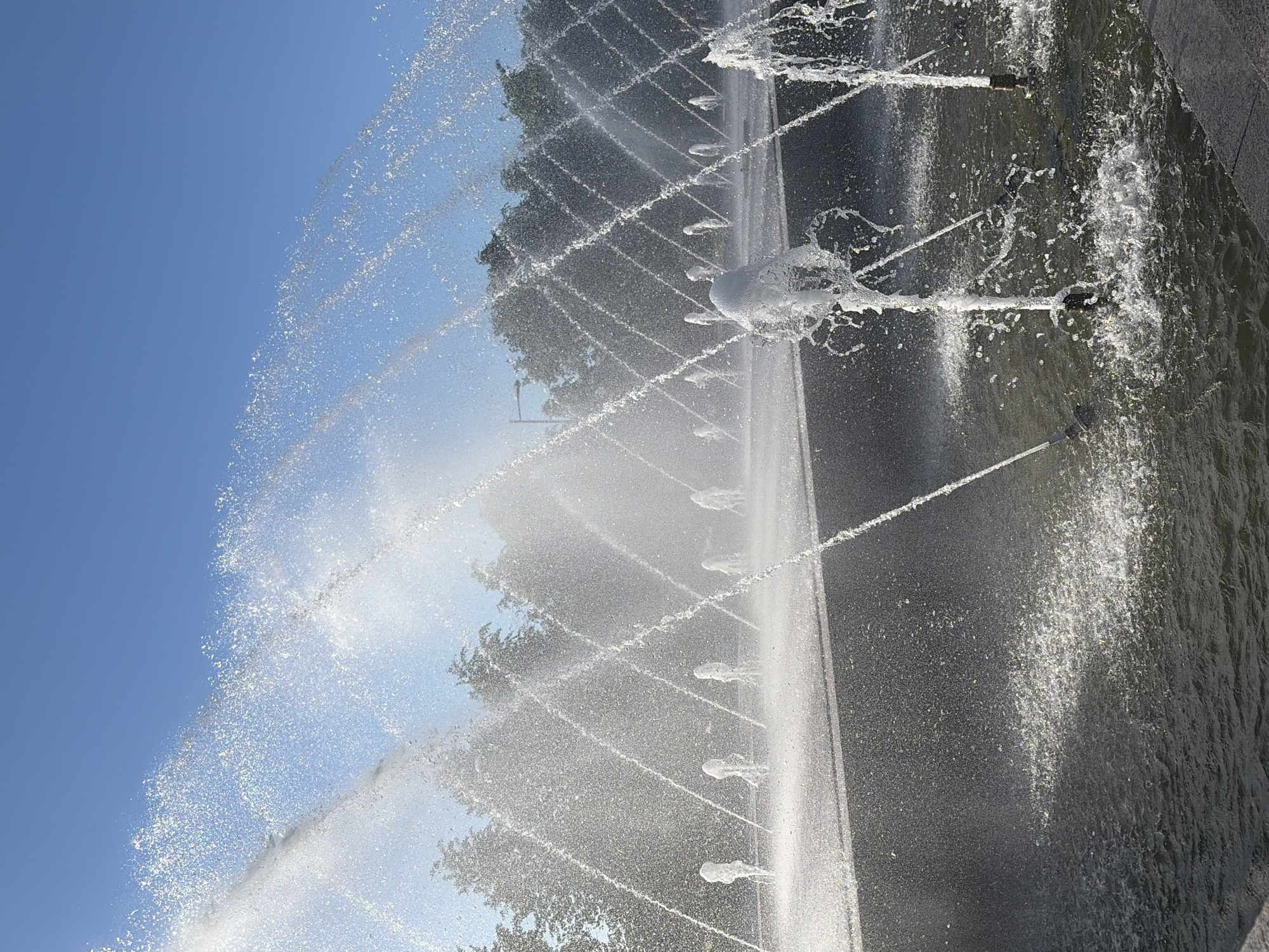 This is a vertical, eye-level shot of a fountain in a public space, likely a park or plaza. The sky is a clear, bright blue, with the sun casting a bright light that creates numerous sparkling water droplets. A line of trees forms the background, with some of their foliage visible through the spray. In the midground, multiple water jets are shooting upwards and outwards, creating a dynamic and textured visual effect. The ground appears to be a dark, flat surface, possibly concrete or stone, with water pooling and reflecting the light. The foreground shows more of the fountain's water jets, with some erupting from the surface of the water. The scene is one of lively and refreshing water activity under a clear sky. The location is identified as Unknown, Russia.