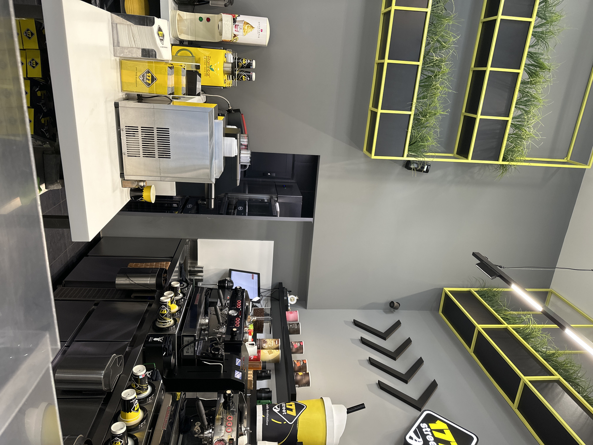 The image shows the interior of a juice bar. The scene is a commercial setting, with a counter featuring an array of equipment: a juicer, a coffee machine, and a refrigerated display case. Yellow and black branded items are noticeable, including juice dispensers and cups bearing the "177 street" logo. The counter surfaces are predominantly white and black. The wall behind the counter is grey and decorated with black arrow-shaped wall fixtures and illuminated black frames containing green plants, adding a decorative element. There are also product displays along the back wall, including coffee cups and possibly other merchandise. The location could be in Serres, Greece, based on the name "177 street" on the signs and cups, although the image alone doesn't conclusively confirm the exact geographic location.