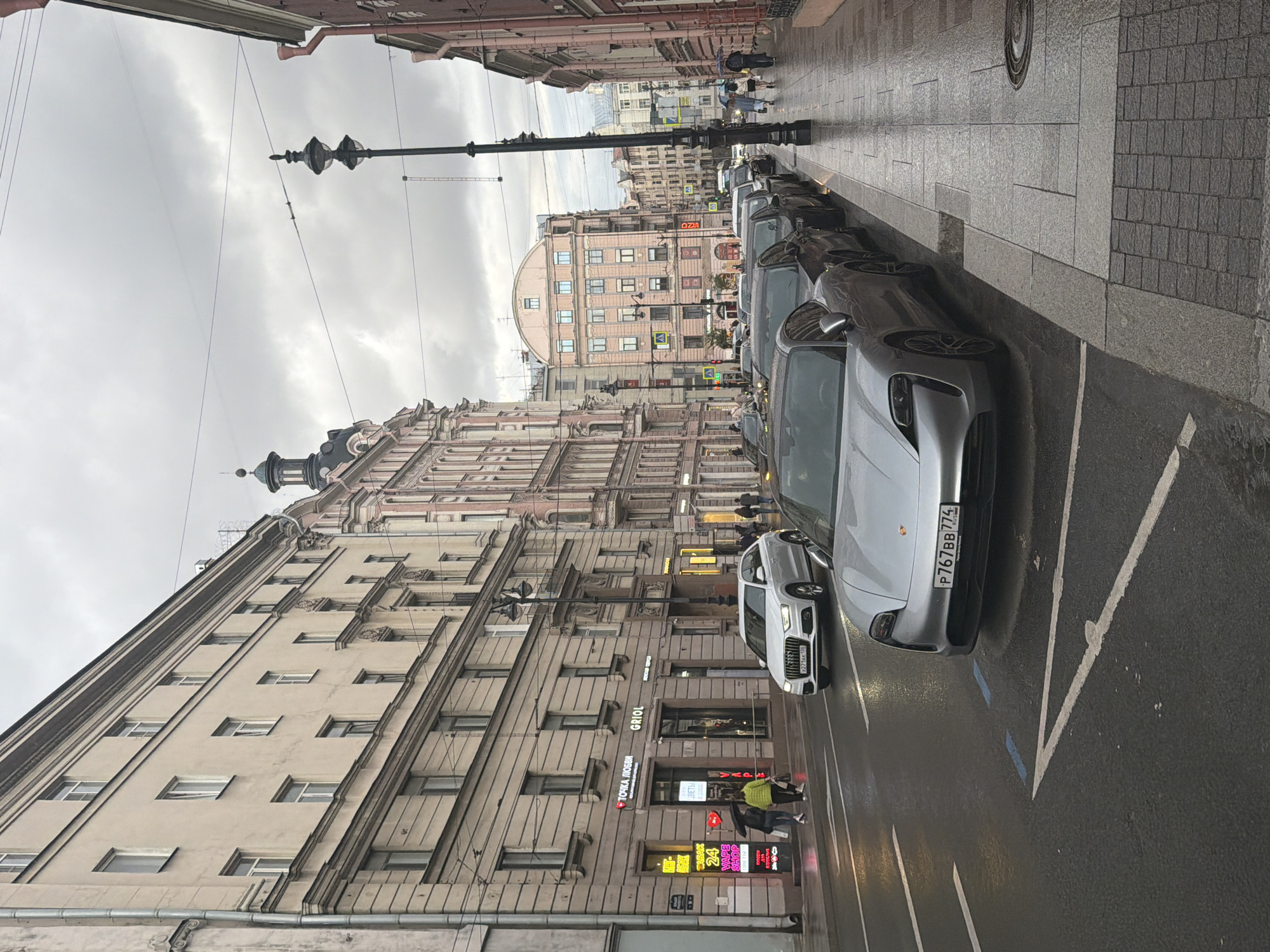 The image shows a city street scene on an overcast day, likely in Saint Petersburg, Russia, given the Cyrillic signage and license plates. A row of parked cars, including a silver Porsche Taycan with license plate P767BB 774 RUS, lines the right side of the street. The street is wet, suggesting recent rain, and reflective. On the left side of the street, there are several buildings with storefronts, including signs for "ТОЧКА ЛЮБВИ" and "GRIOL". A few pedestrians are visible, some with umbrellas, walking on the sidewalk. The architecture suggests an older European city with multi-story buildings featuring ornate details. A lamppost stands on the right side of the sidewalk.