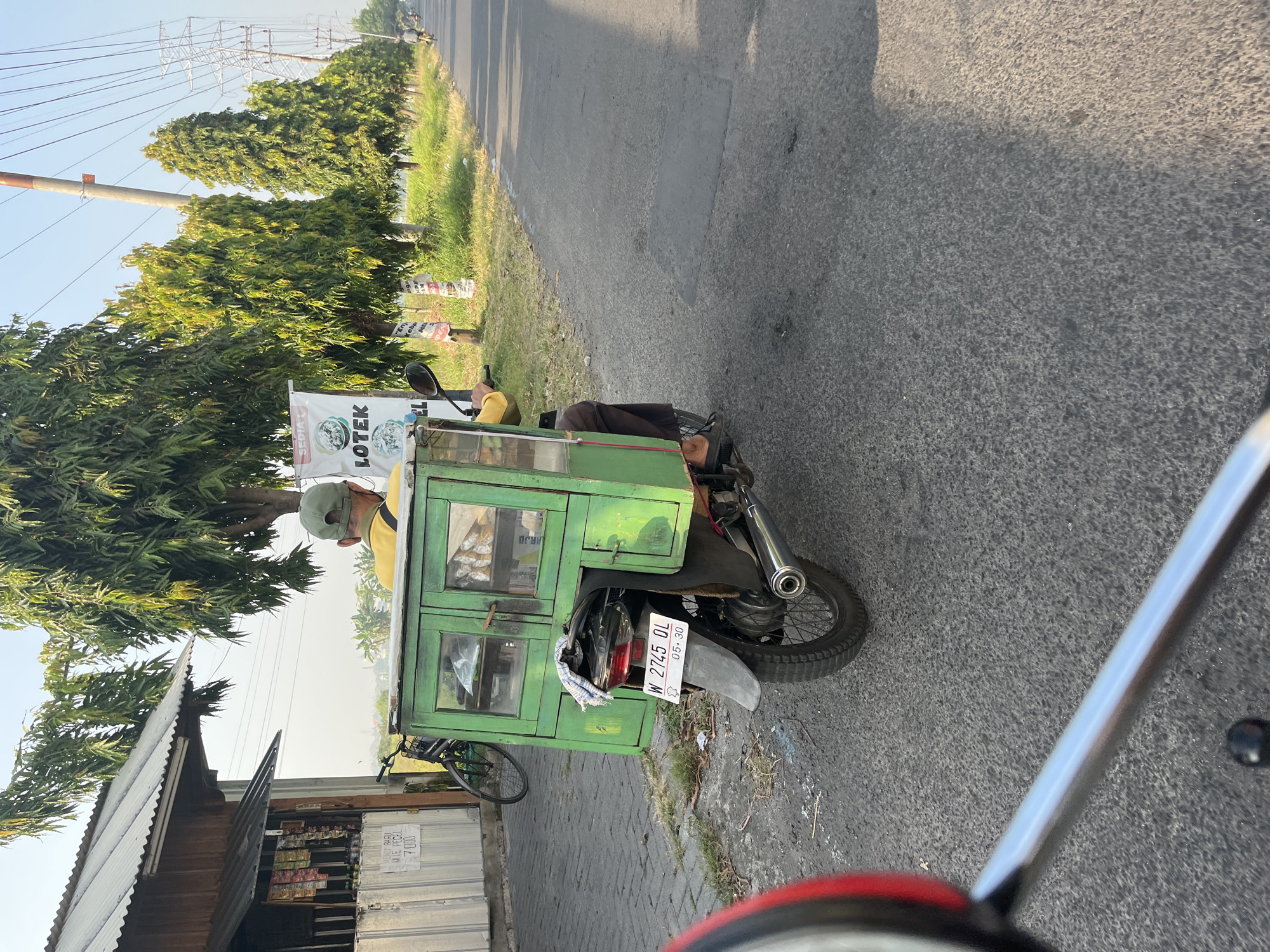 A man wearing a yellow shirt and a green cap rides a motorcycle with a green wooden food cart attached to the back. The cart has glass doors displaying food items and a license plate that reads "W 2745 QL". The motorcycle is on a paved road in what appears to be a semi-rural area, with trees lining the road and a small building with goods for sale on the left. In the background, power lines and transmission towers are visible. The scene is set in Karangtanjung, Indonesia, during daylight hours.