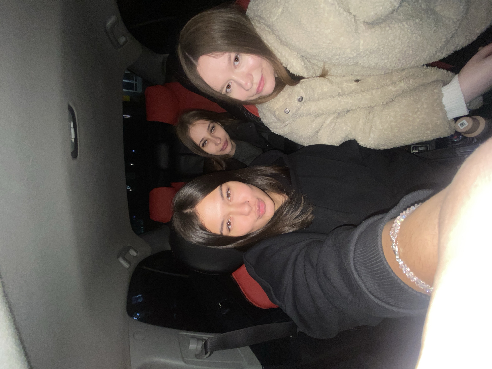 Three young women are inside a vehicle, possibly a car, likely taking a selfie. The interior of the vehicle is dark with red seats. The woman in the foreground is wearing a black hooded sweater and a beaded bracelet. The woman in the middle has shoulder-length brown hair and is wearing a dark top. The woman in the background is wearing a fluffy beige jacket. The location is likely in the city of Chaikovsky, Russia, based on the image request.