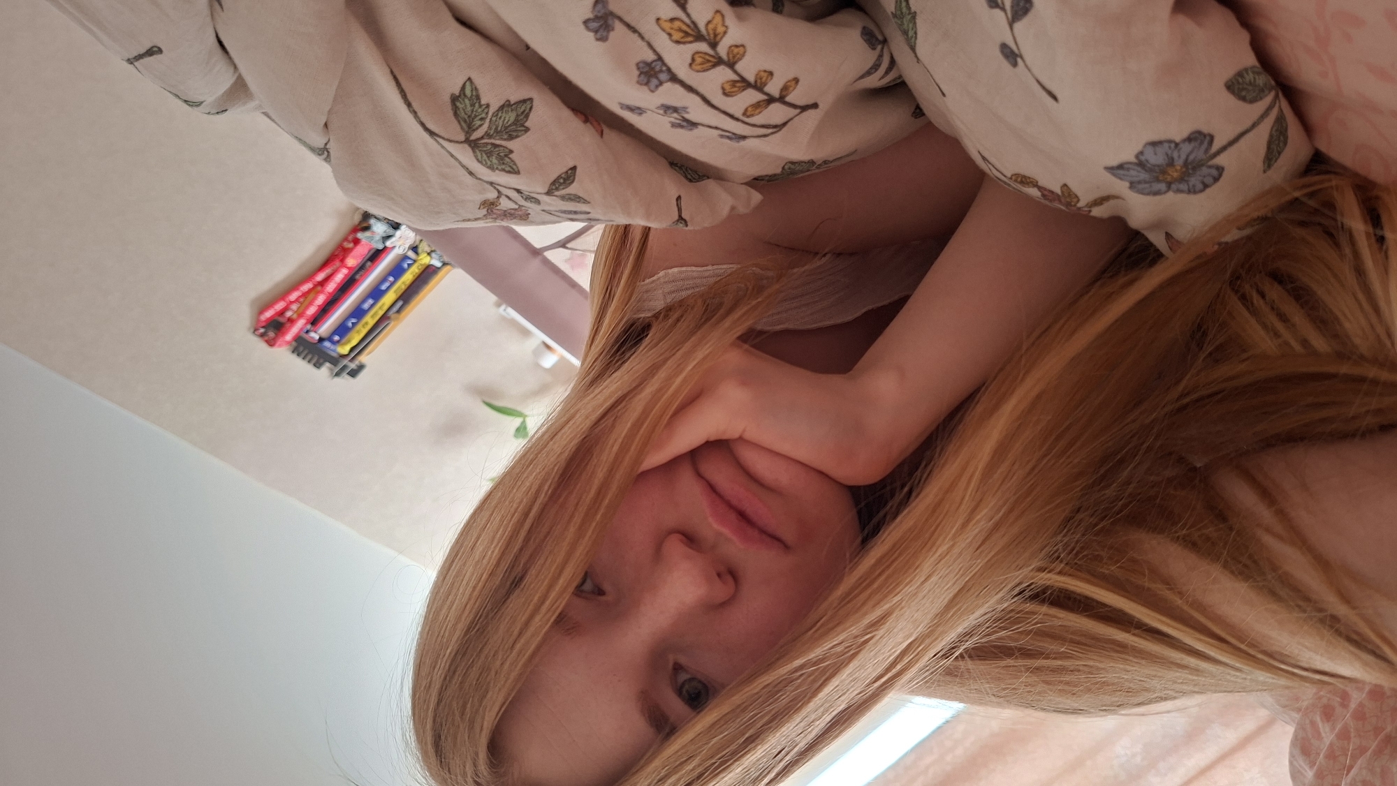 A young, light-skinned person with long, straight reddish-blonde hair is visible from the chest up. They are holding their cheek with their left hand, appearing partially covered by bedding with a light floral pattern. In the background, there's a glimpse of a wall and a row of colored pencils or markers. The location is inferred to be the city of Chaikovsky, Russia.