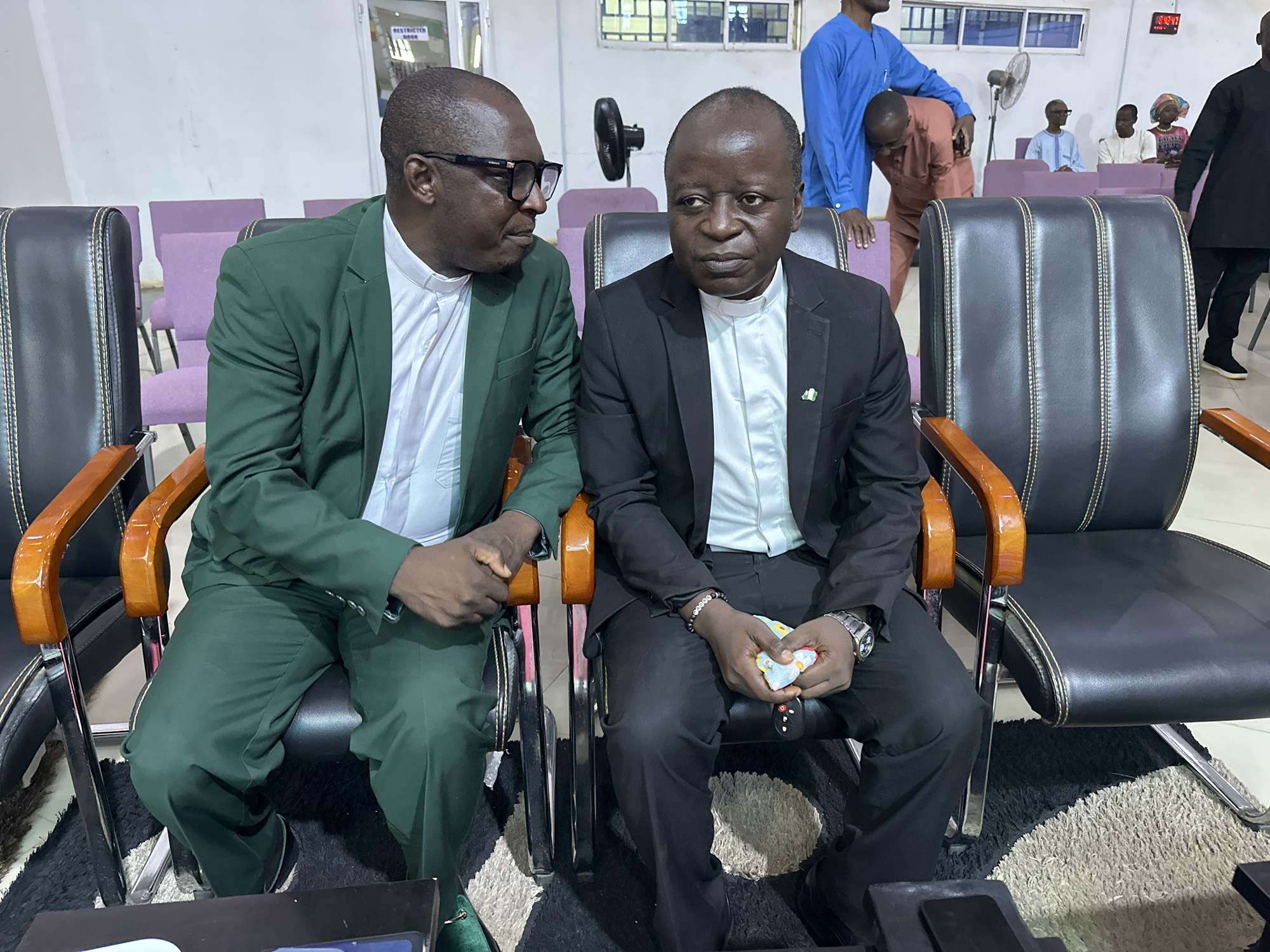In Goni Gara, Nigeria, two men in clerical attire are seated in black leather chairs in what appears to be a meeting room or hall. The man on the left, wearing a green suit over a white clerical collar, has glasses and is looking towards the man on the right. The man on the right, dressed in a black suit with a white clerical collar, is looking forward. Both are seated next to each other, with their hands visible. The background shows other individuals and rows of purple chairs, suggesting a gathering. A large fan is visible in the distance. The lighting is bright, indicating an indoor setting.