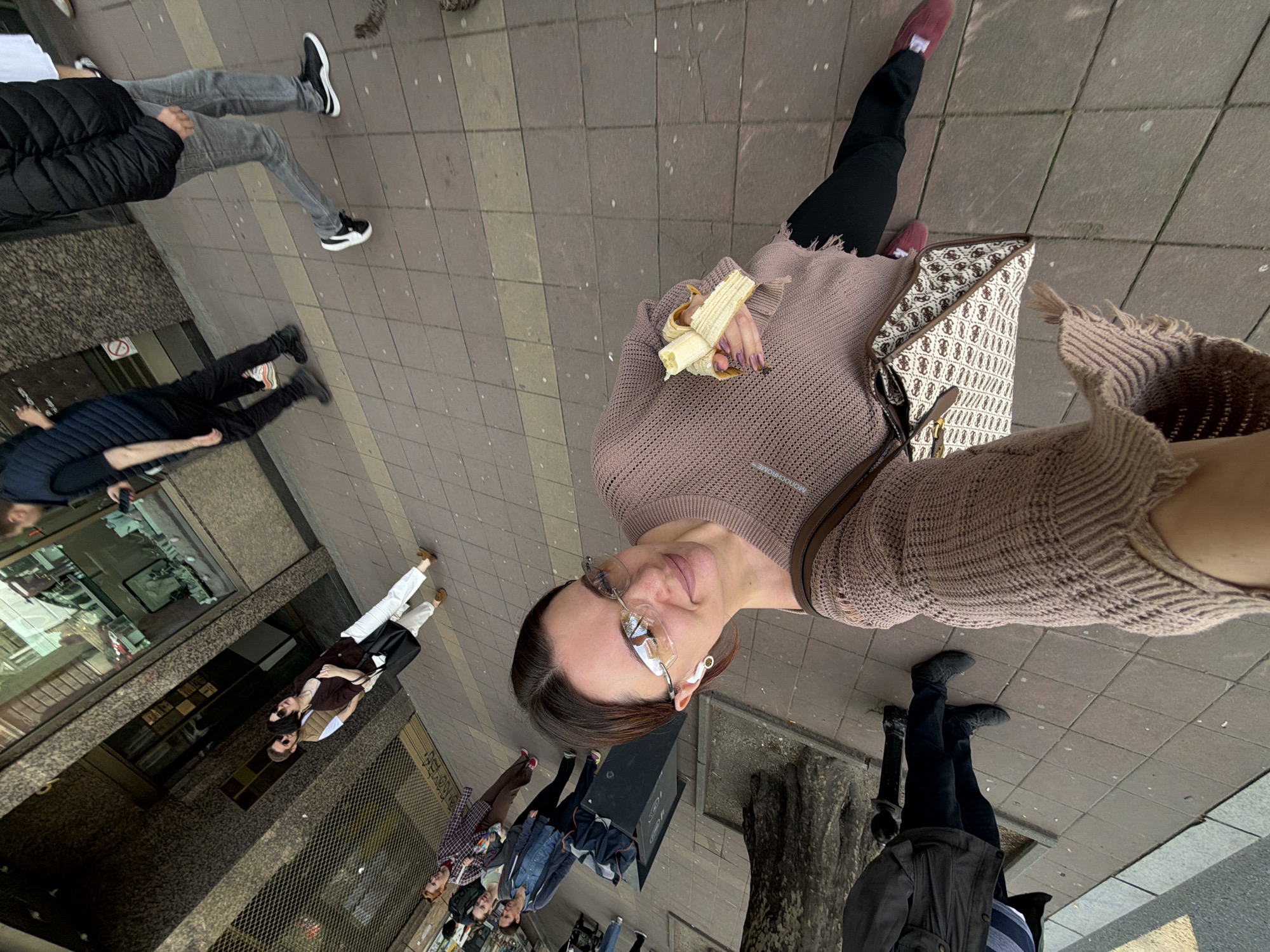 The image is an overhead selfie taken on a street in Belgrade, Serbia. The photographer is a woman with glasses, a brown sweater, and a designer handbag, holding a half-eaten banana. She is standing on a brick sidewalk. Around her are other pedestrians, including a couple in the reflection of a store window, and others walking on the sidewalk. Some are wearing casual clothes. In the background is a tree next to a bin. The sidewalk is made of gray and brown bricks.