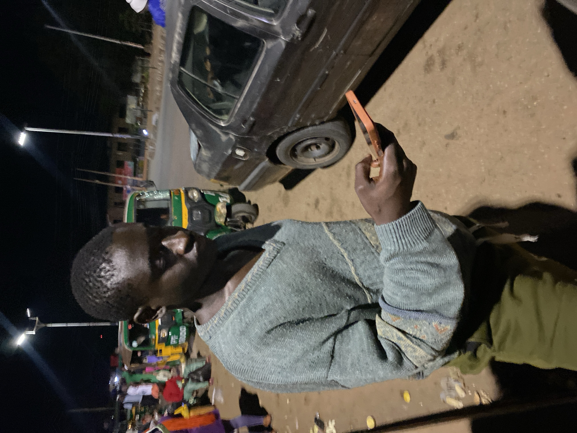 An African male, appearing to be in his late teens or early twenties, stands outdoors at night in what appears to be a public area in Bukuru, Nigeria. He is wearing a light blue, V-neck sweater over olive green sweatpants. His attention is focused on an orange-cased smartphone he holds in his right hand.
The background suggests a busy roadside or marketplace. To the left, several auto-rickshaws, known locally as "keke napeps," are visible, their green and yellow bodies illuminated by streetlights. Some people, dressed in casual attire, are milling around in the background. A dark-colored sedan is parked to the right of the frame. Overhead streetlights provide the primary illumination, casting long shadows on the dusty ground, indicating a clear, dry night. The general ambiance suggests a lively but somewhat unkempt urban environment.