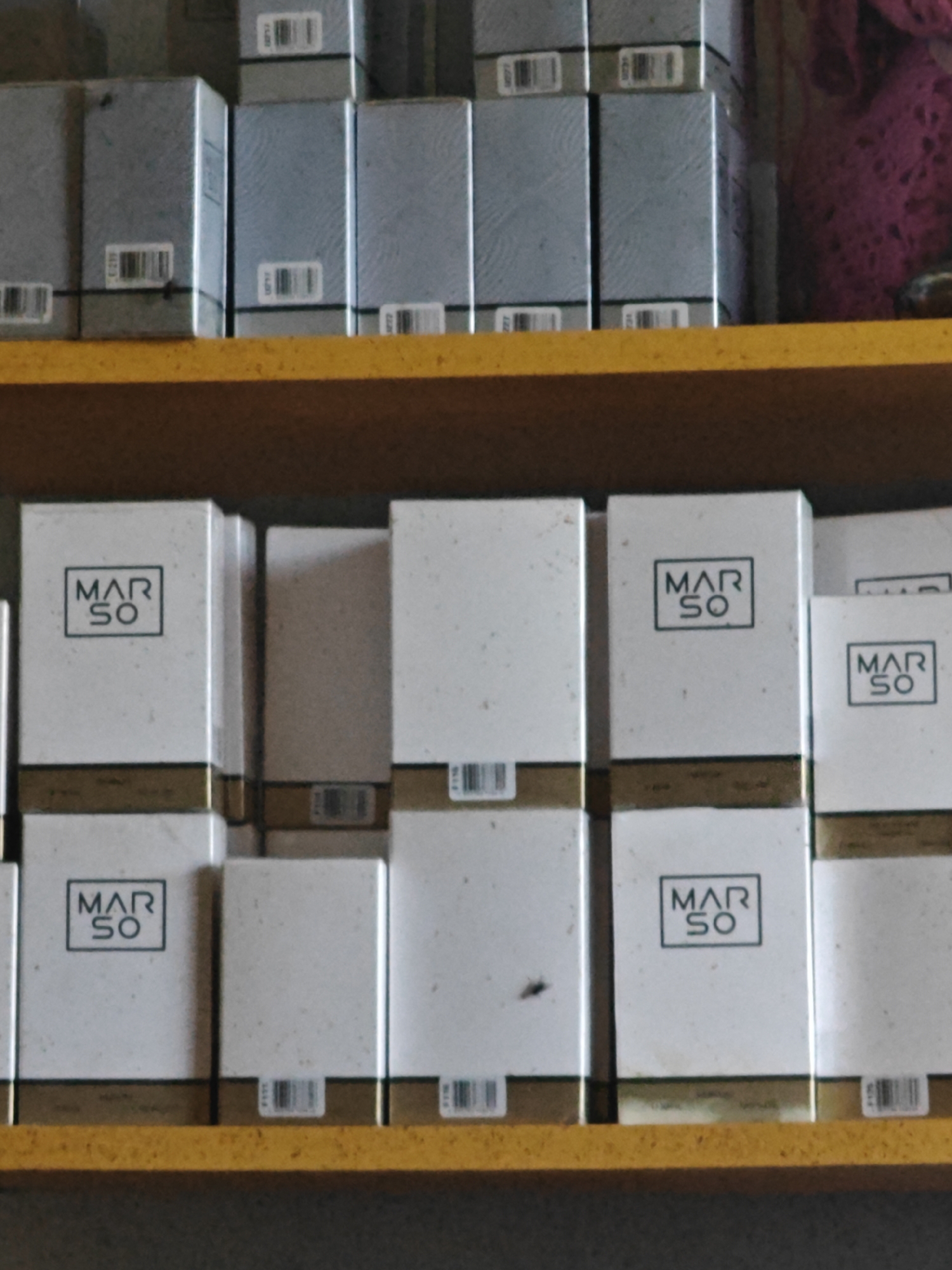 The image shows a shelf containing numerous white boxes with the "MAR SO" logo. These boxes appear to be perfume or cosmetic products. The boxes are stacked in rows, with some of the boxes on the top shelf being a light blue-gray color and also displaying barcodes. The shelf is made of yellow wood. The background is dark and appears to be a wall. There are no people or animals visible in the image. The image quality is moderate, with some graininess and blurriness, suggesting it was taken in low light conditions or with a lower resolution camera. The scene is indoors. There is no information in the image that specifically indicates the location is Magadan, Russia.