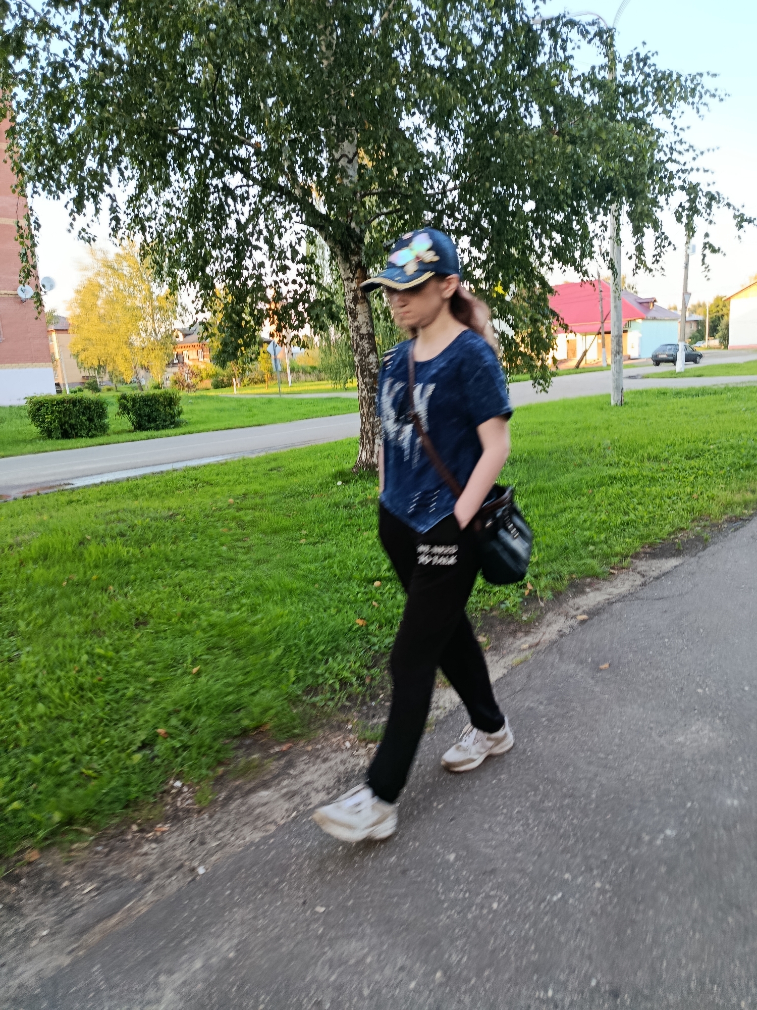 A person walks on a paved path next to a grassy area. The person is wearing a blue t-shirt, black pants, a baseball cap, and white sneakers. They are carrying a dark-colored bag over their shoulder. In the background, there is a street with a car, houses with red and blue roofs, trees, and bushes. The sky is visible above. The scene appears to be in the city of Vereya, Russia.