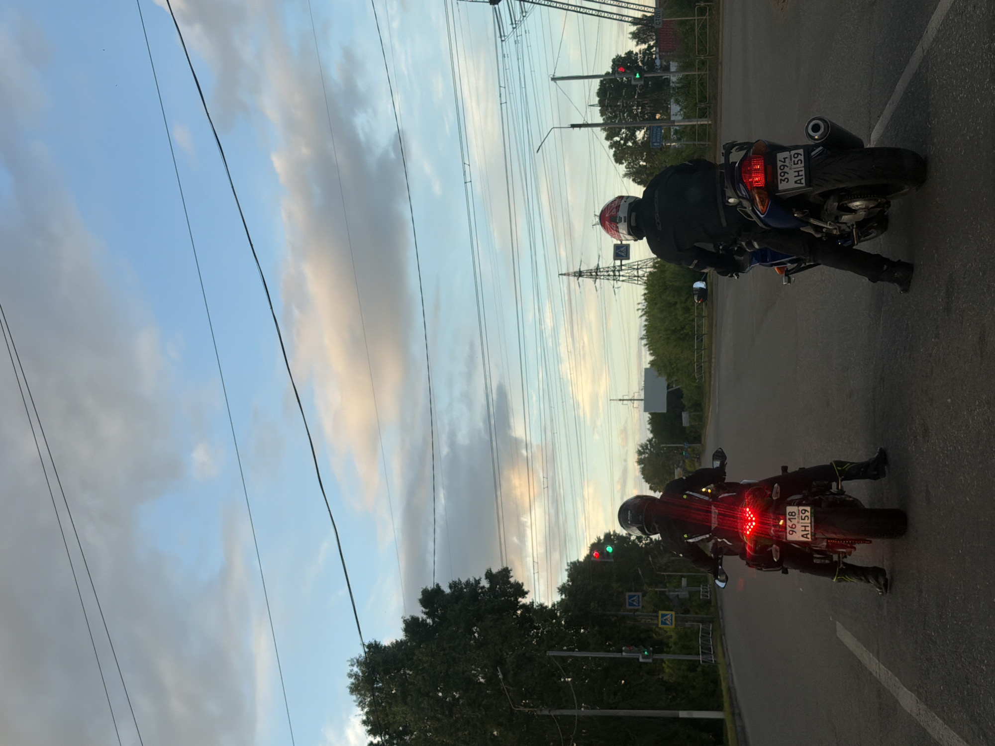 The image depicts two motorcycles, each with a single rider, stopped at a traffic light in Chaikovsky, Russia. Both motorcycles are facing away from the camera. The motorcycle on the right is blue and white with a license plate that reads "3994 AH 59." The rider is wearing a black jacket and a red and white helmet. The motorcycle on the left is black and has a license plate reading "9618 AH 59." The rider is wearing a black helmet and jacket. Both motorcycles have illuminated taillights. The background includes trees, power lines, and a traffic light with a green signal. The sky is cloudy with hues of blue and gray.
