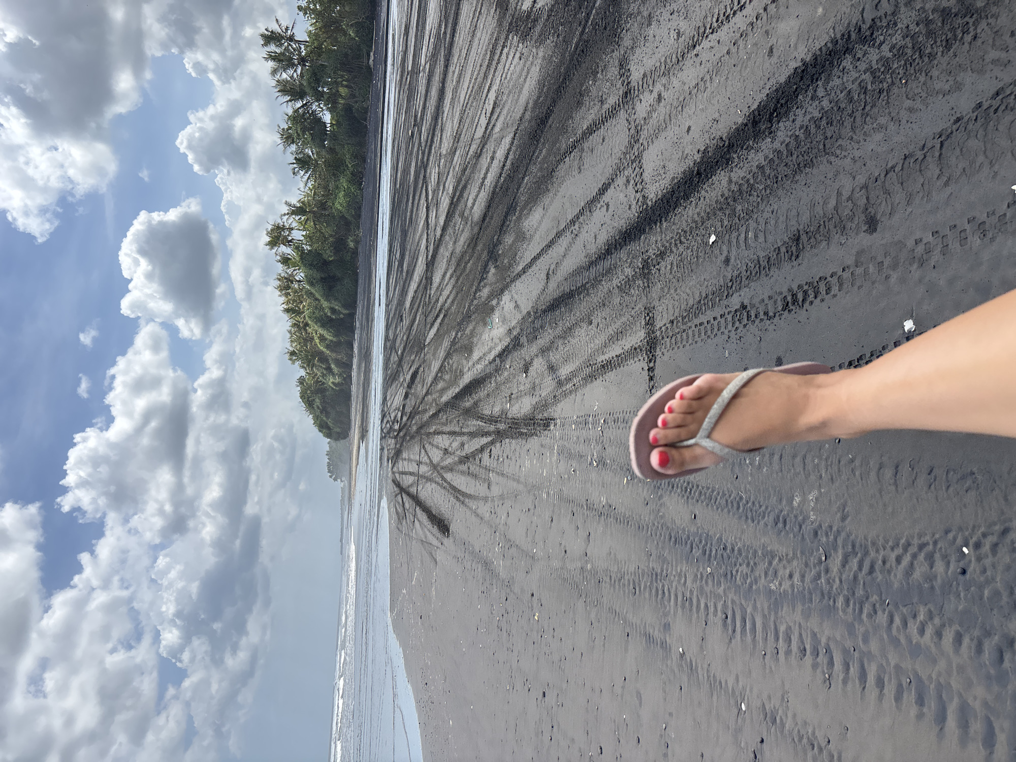 The image shows a beach scene in Kerambitan, Indonesia. The beach has black sand marked with tire tracks. The ocean meets the sand on the left of the frame. Vegetation lines the beach in the background. The sky is visible above with a combination of blue sky and white clouds. A fair-skinned person's foot wearing a silver sandal with red painted toenails is visible on the right side of the frame.