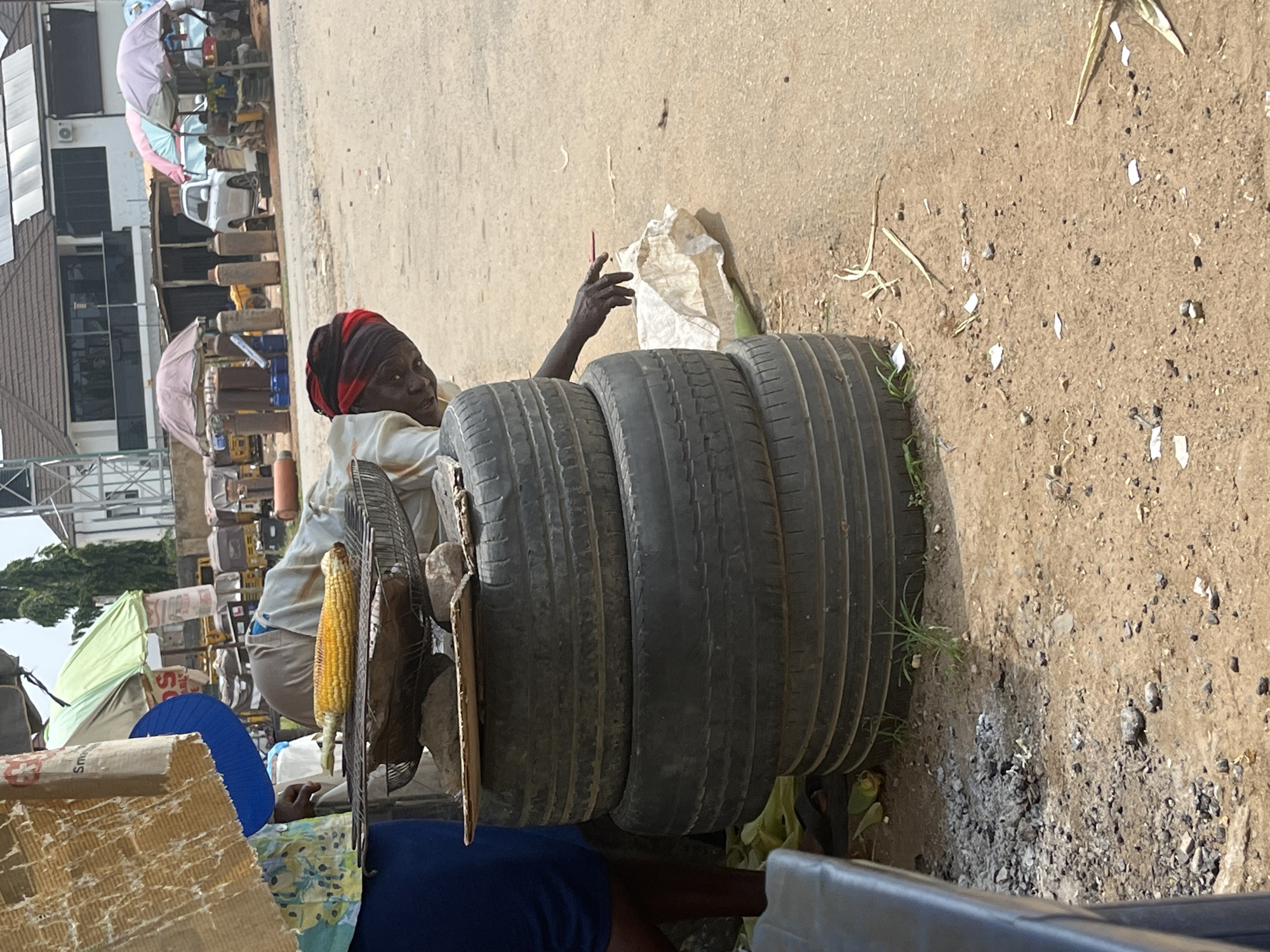 The image captures a street scene in Abuja, Nigeria, with a focus on a street vendor preparing food. In the foreground, three stacked tires serve as a makeshift stand. On top of the tires, a metal grill holds a partially roasted ear of corn. A person, wearing a red headwrap and a light-colored shirt, is leaning over the grill, attending to the corn. Their right arm is extended, and their hand is reaching out towards the ground, where a white plastic bag and some dry vegetation are visible. To the left, a person wearing a blue skirt is partially visible. In the background, there are market stalls with colorful umbrellas, various vehicles, and buildings, suggesting a bustling urban environment. The ground is a mixture of dirt and gravel, with some scattered debris.