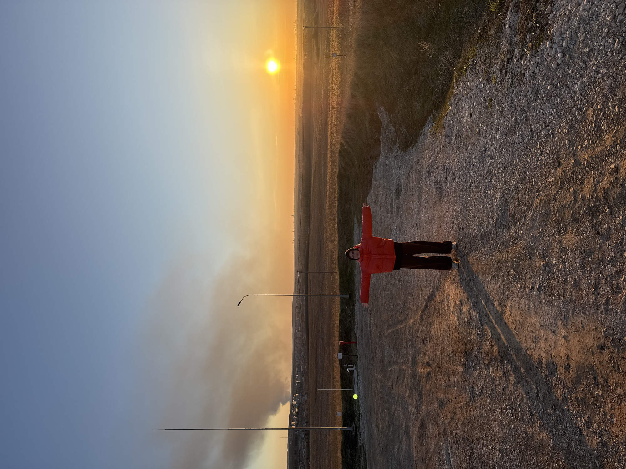 A photograph shows a person standing on a gravel path with arms outstretched at sunset in Лобаново, Russia. The sky transitions from blue to orange near the horizon, where the sun is visible. A field stretches into the distance, dotted with utility poles and what appears to be a cloud of smoke or dust near the base of the picture. The person is wearing a red jacket and dark pants. The path is uneven with gravel, and shadows are long, suggesting the low angle of the sun.