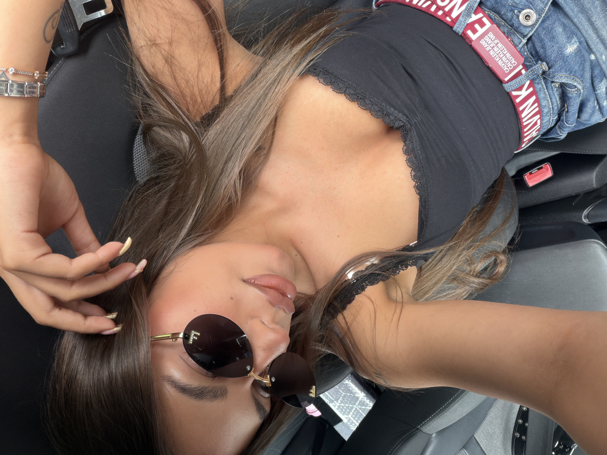 A young woman with long, brown hair is seen from a high-angle, close-up perspective, looking down at the camera. She is wearing dark sunglasses with "F" logos on the lenses, a black lace-trimmed tank top, and denim jeans with a red Calvin Klein belt. Her right hand is resting on her head, with long, manicured nails. She appears to be in a car, indicated by the presence of a seatbelt buckle and car seat material. The lighting suggests it is daytime, possibly outdoors or in a well-lit interior. There is no visible indication of her location within the city of Burgas, Bulgaria.