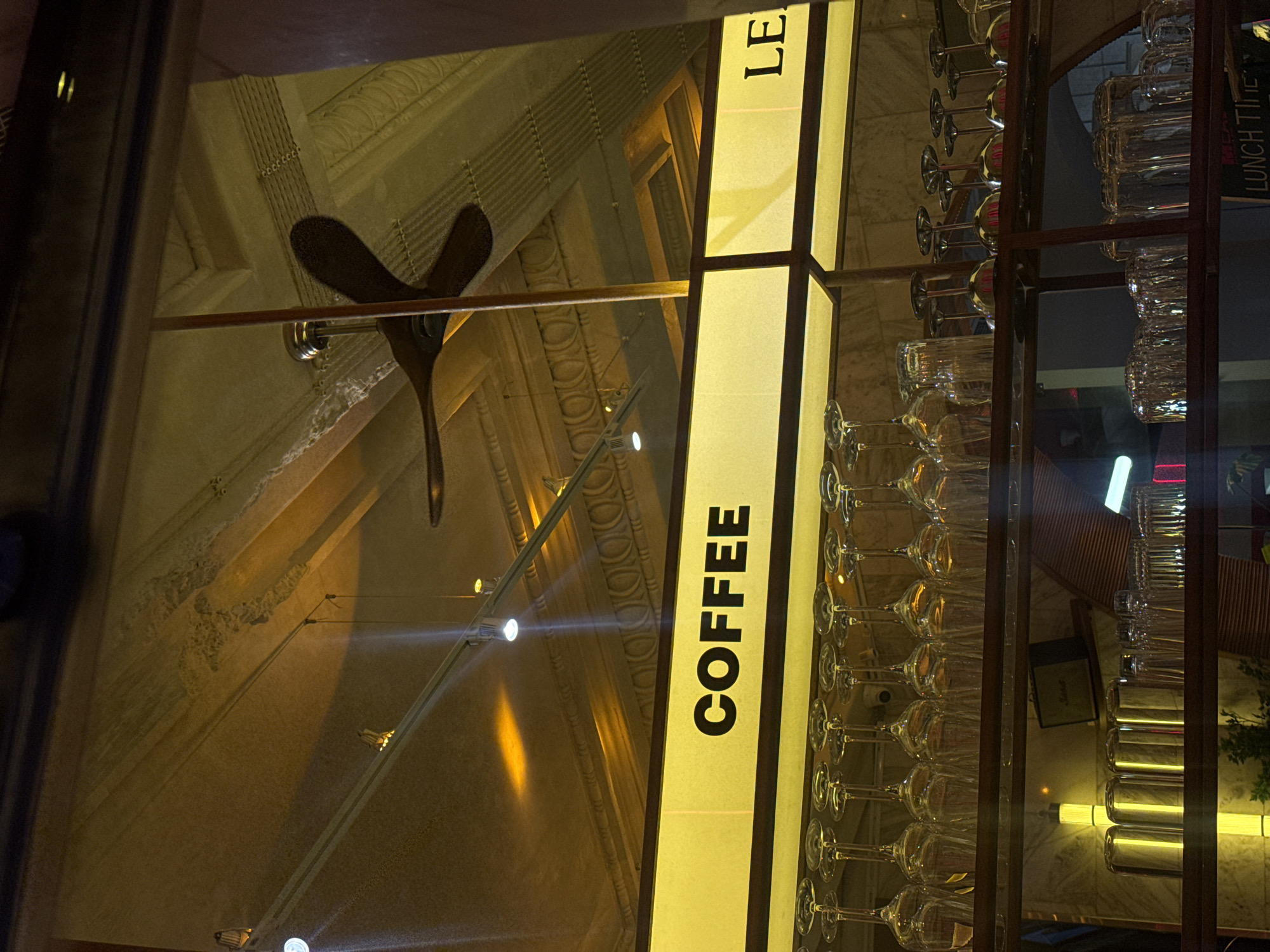 The image depicts an interior view of Coffee LE Street Bar, located at 2 Abovyan Street, Yerevan, Armenia. The perspective is looking upwards and across a service area, possibly from a patron's viewpoint or through a window. The upper section of the frame shows a high ceiling with elaborate, light-colored decorative moldings and cornices, illuminated by warm, yellow-orange ambient light. Linear track lighting fixtures are mounted on the ceiling, casting distinct light beams. A dark, three-bladed ceiling fan is suspended near the center-right. Below the ceiling structure, a prominent, yellow-backlit sign displays the word "COFFEE" in black capital letters. To its right, an adjacent sign shows the partial letters "LE". Below these signs, a metal shelving unit, likely part of a bar, is visible. The top shelf holds multiple clear stemmed wine glasses hanging upside down. The middle shelf contains various upright clear drinking glasses, including tumblers, alongside several silver-toned, conical bar tools or shakers. A security camera (CCTV) is mounted above the top shelf, centrally positioned. A dark Marshall brand speaker rests on a lower shelf in the background. The bottom shelf features more upright clear drinking glasses, and a partial sign reading "LUNCH TI" is visible at the extreme bottom right corner. No individuals are present in the scene.