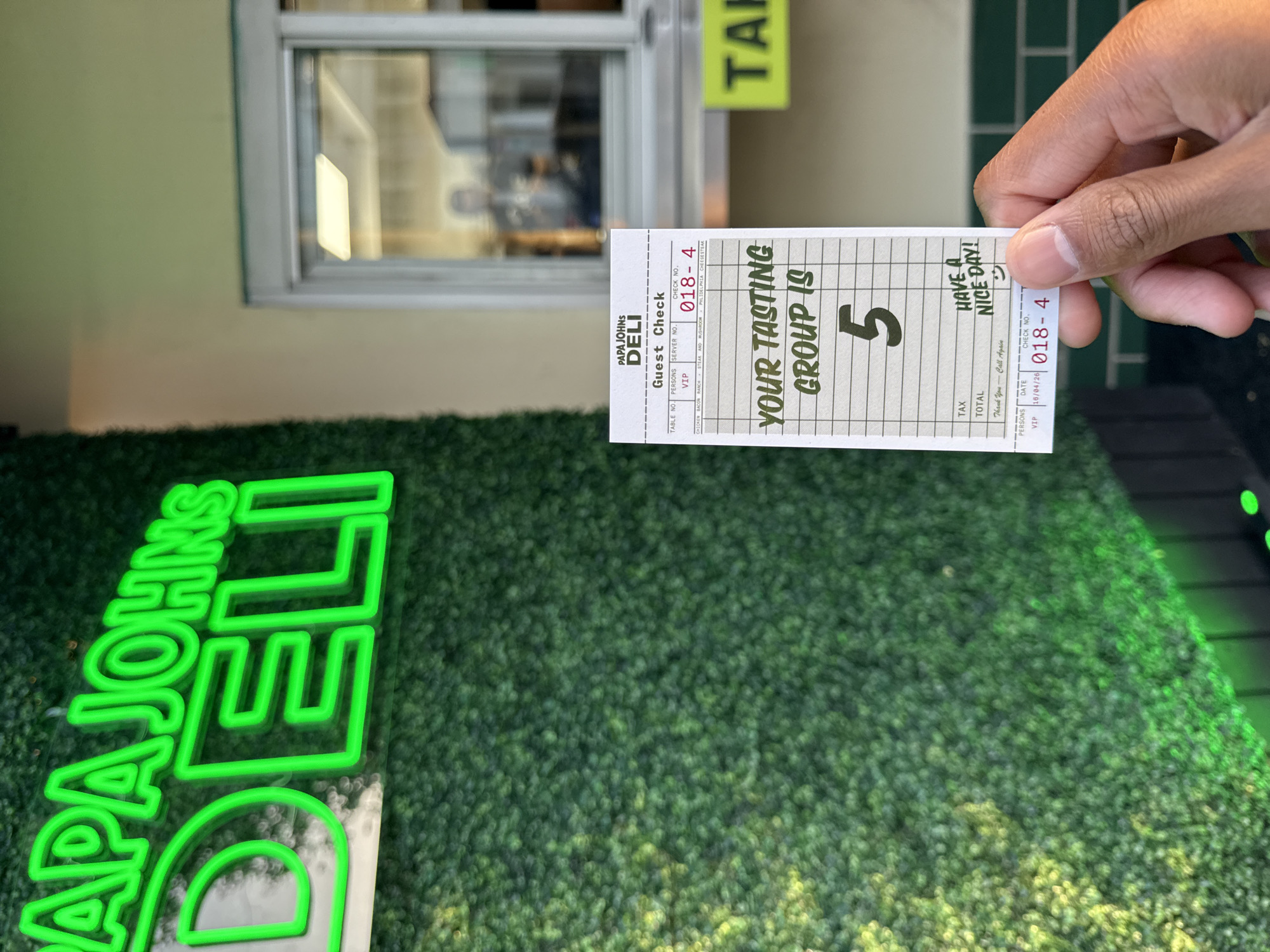 A medium-angle shot captures a person's hand holding a guest check from "PAPA JOHNS DELI" in what appears to be an outdoor or semi-outdoor setting. The guest check is the central focus, displaying the establishment's name, "Guest Check," and "CHECK NO. 018-4." Prominently written on the check in larger font is "YOUR TASTING GROUP IS 5." Other details on the check include "CHICKEN BACON," "PHLIDELPHIA CHEESESTEAR" (with a visible misspelling of Philadelphia), "Thank You - Call Again," "HAVE A NICE DAY!", and the date "16/04/26." The hand holding the check is lightly tanned with visible knuckles.
In the foreground and left, a bright green neon sign spells out "PAPA JOHNS DELI," positioned against a vibrant green artificial foliage wall that covers much of the ground and extends vertically. This green wall forms a textured backdrop for the scene. In the background, a window reflects a blurry interior or exterior view, suggesting a building facade. Above the window, part of a yellow sign with dark green text, reading "TAH," is visible. The lighting suggests it is daytime, although the neon sign is illuminated. The overall scene indicates a casual food service environment in Los Angeles, United States of America, likely related to a tasting event or group dining experience.