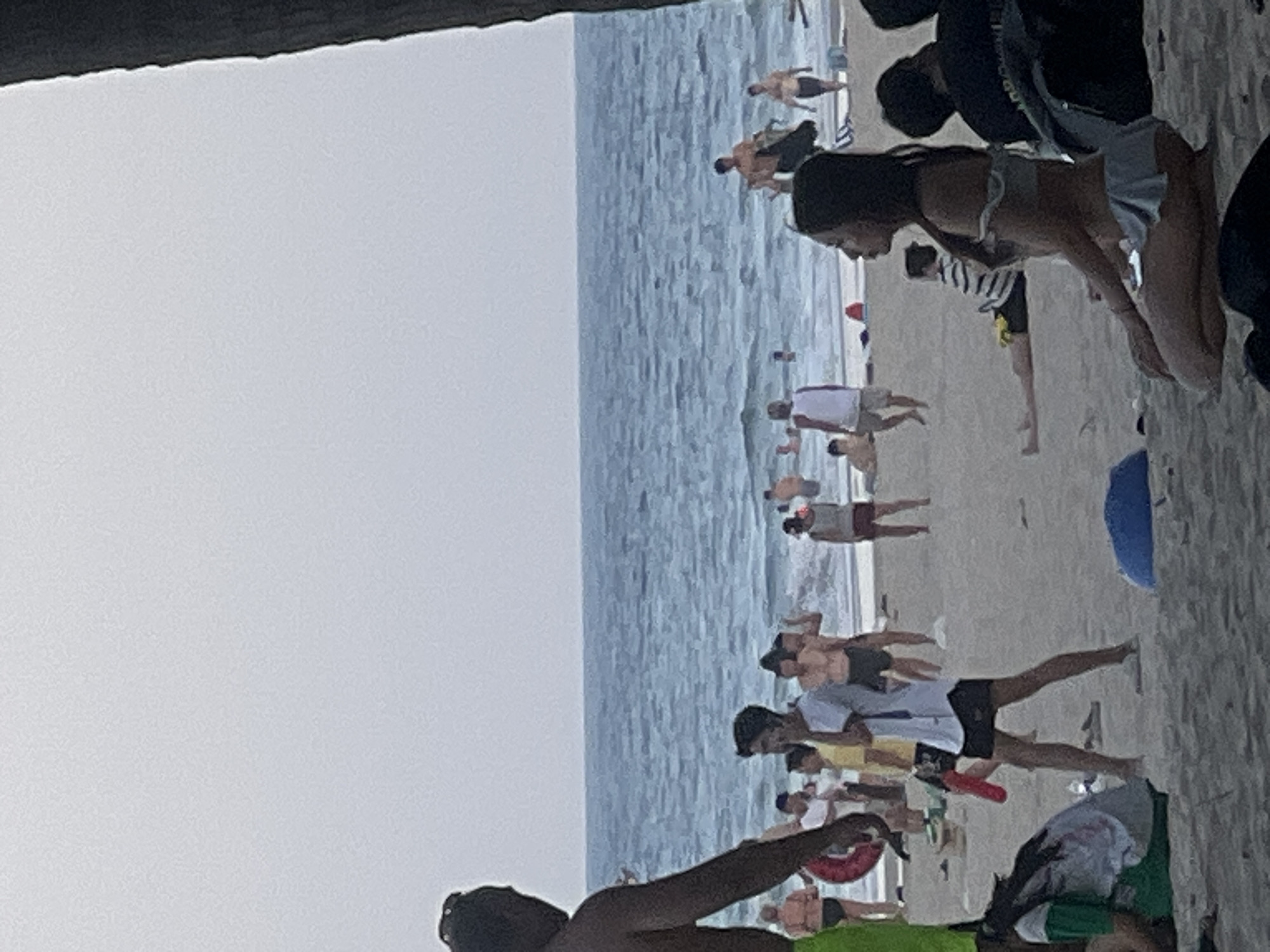 The image depicts a crowded beach scene, likely in Đà Nẵng, Vietnam. Numerous people are present on the sandy shore, with the ocean and waves visible in the background. The sky is overcast, suggesting it might be a cloudy day or late afternoon. The people are dressed in beachwear, and many appear to be walking, standing, or engaging in recreational activities. Some individuals are in the water, enjoying the waves.
In the foreground, a person's arm and shoulder are visible, along with part of their head, suggesting they are facing away from the camera. Further into the scene, a woman in a light-colored bikini is kneeling. Other people are scattered across the beach, some in groups and others individually. There are various objects on the sand, including what appears to be a blue inflatable ball or raft and a red inflatable ring. The atmosphere seems casual and relaxed, typical of a beach day. The presence of numerous people suggests it's a popular spot. The image is slightly blurry and tilted, making precise details difficult to discern, but the overall impression is of a lively beach with many visitors enjoying the seaside. There is no visible text in the image.