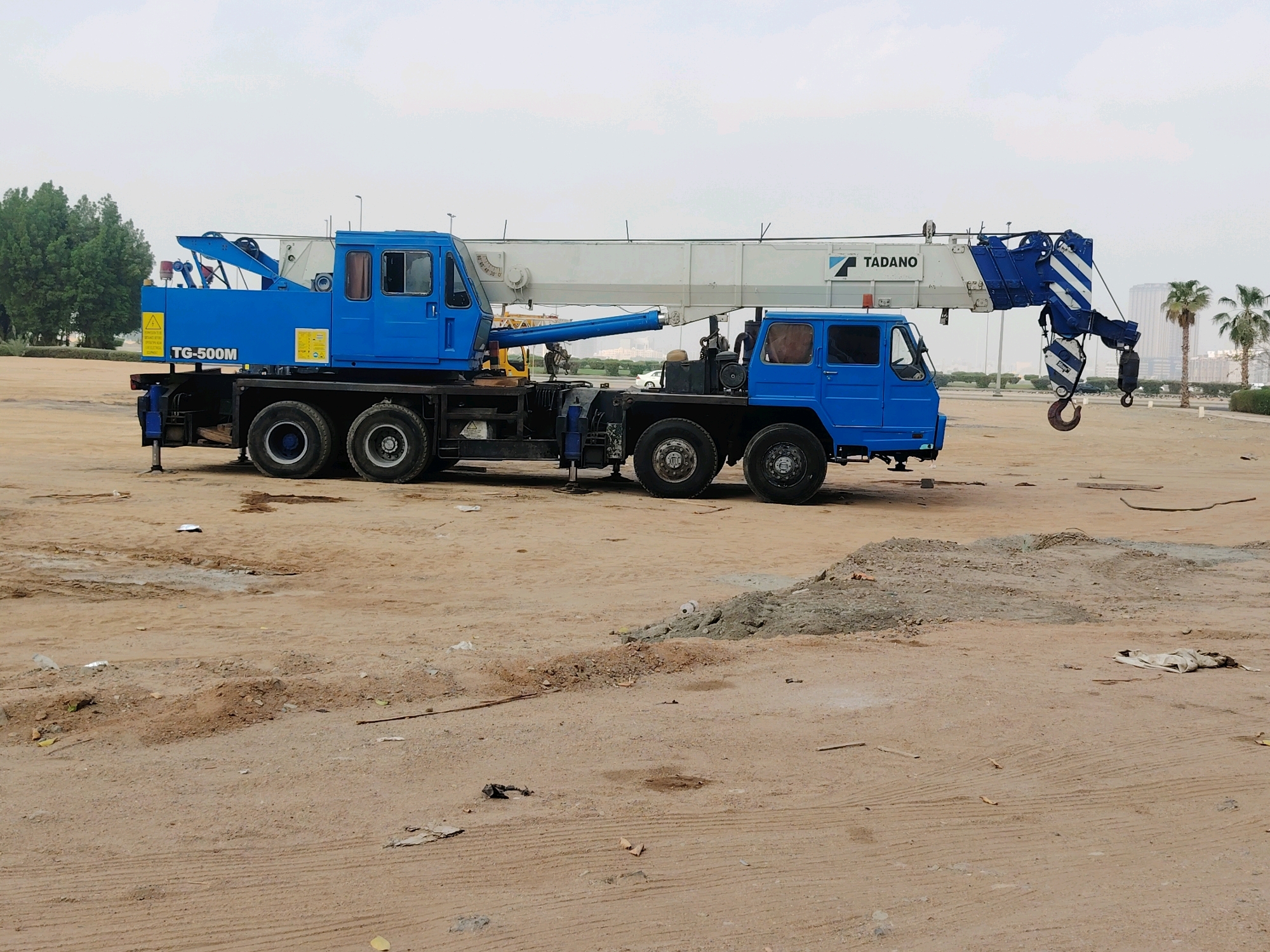 An exterior, eye-level, full shot captures a blue Tadano TG-500M truck crane parked on a dirt lot in Jeddah, Saudi Arabia. The crane is the primary subject, taking up most of the frame, with its long white and blue boom extended and angled slightly upwards. The crane's cab, also blue, is visible on the left, and a second, smaller blue cab is situated on the right side of the truck chassis. The crane is equipped with outriggers extended downwards.
The environment is an undeveloped construction or staging area. The ground is composed of dry, sandy soil with some scattered debris, including pieces of concrete and possibly construction materials. In the background, a line of green trees runs across the middle ground, and beyond that, the distinctive skyline of Jeddah, with its modern buildings and palm trees, is visible under a hazy, overcast sky. The overall impression is one of an industrial or construction site in a semi-urban environment. No people are visible in the image.