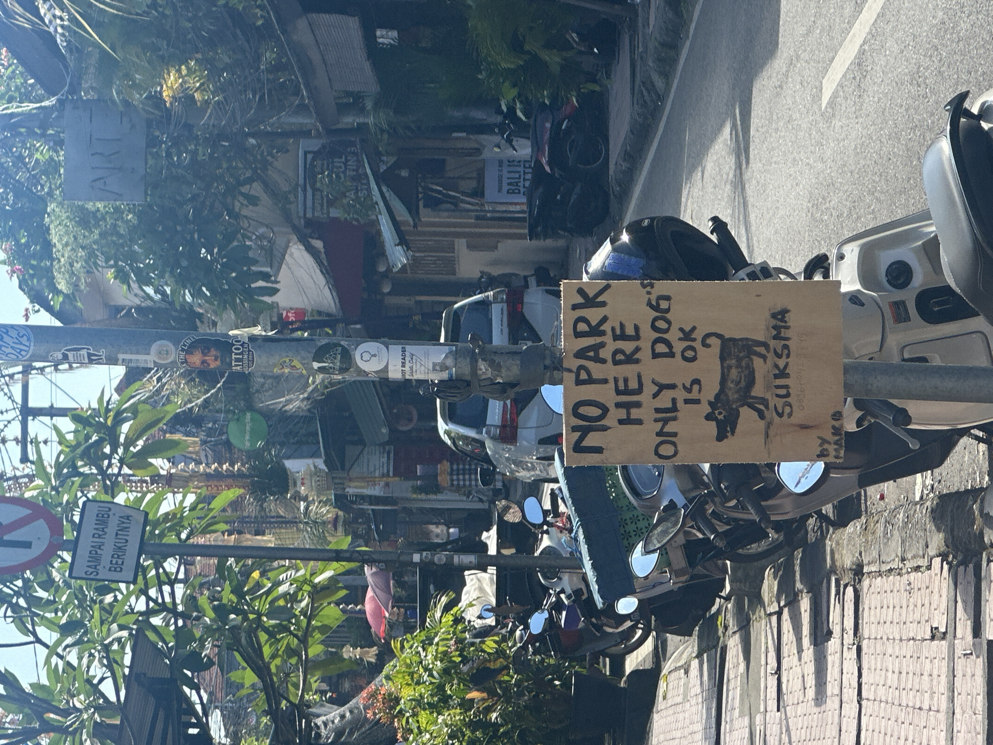 The image captures a street scene in Ubud District, Indonesia, under a bright, sunny sky. In the foreground, a handmade sign on a wooden plank is attached to a pole. The sign, written in black marker, reads "NO PARK HERE ONLY DOG IS OK" with a drawing of a dog below. It also has "by MAKO" written at the bottom. Several motorbikes are parked along the curb.
Behind the motorbikes, a light-colored car is visible, and further down the street, more buildings and trees line the road. Various other signs are attached to the pole in the foreground, including one with a red circle and a diagonal line over a symbol, and another in Indonesian text. One sign, partially obscured, appears to have the word "ART" on it. The overall impression is a bustling street in a tropical location, with a touch of humor provided by the sign.