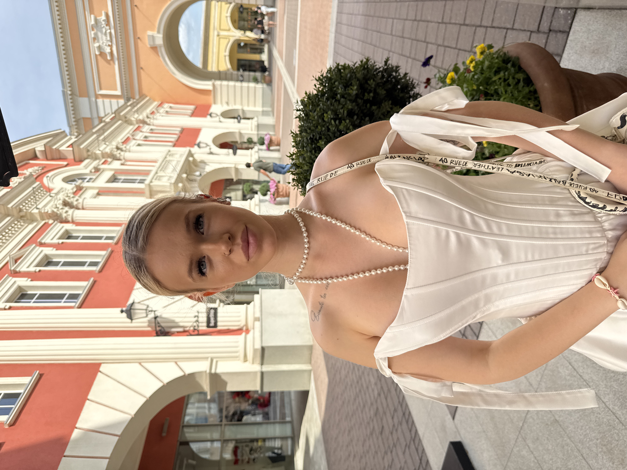 The image captures a young woman with blonde hair tied back, standing outdoors in front of a European-style building with red and orange facades. She is wearing a white, off-the-shoulder corset-style top with ribbons tied at the shoulders, a pearl necklace, and a bracelet adorned with shells. Her handbag, with a patterned strap featuring text, is slung over her shoulder. The background shows architectural details like arches, columns, and windows, with glimpses of other people in the distance. Potted plants with yellow and pink flowers are visible on either side of the woman. The ground is paved with bricks and tiles. The scene appears to be a shopping arcade or a similar public space in a city. There is no specific indication in the image that places this scene in Saint Petersburg, Russia.