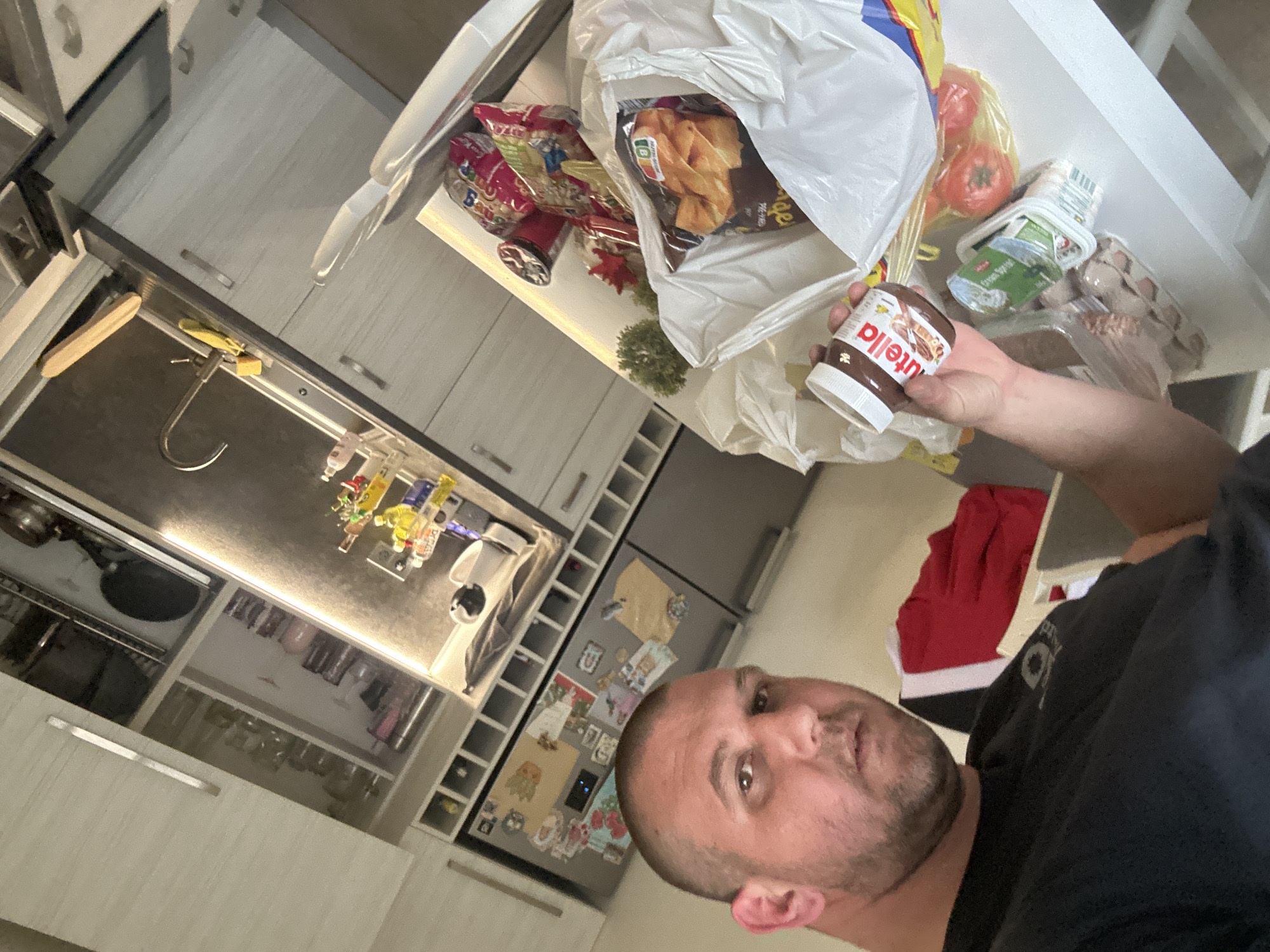 A man is holding a jar of Nutella in his right hand in a kitchen. A white plastic shopping bag is on the counter with groceries including tomatoes, eggs, lettuce, chips, and cereal. The kitchen has white cabinets, a stainless steel sink, and a refrigerator with magnets. The location is a kitchen interior, presumed to be in Sofia, Bulgaria.