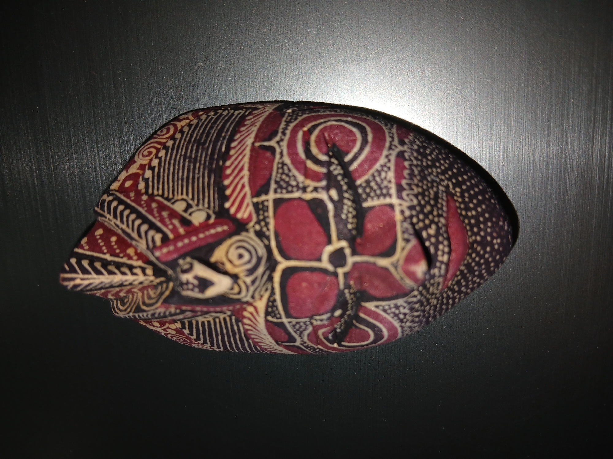 A patterned head covering is placed on a dark gray, possibly metal, surface. The head covering has a rounded, elongated shape, tapering to a point at one end. It features a complex batik-like pattern in shades of dark red, beige, and dark purple. The design includes floral motifs, geometric shapes, and curvilinear lines. The lighting is uneven, creating highlights and shadows across the surface. The context or location in Bengaluru, India, is not directly apparent from the image.