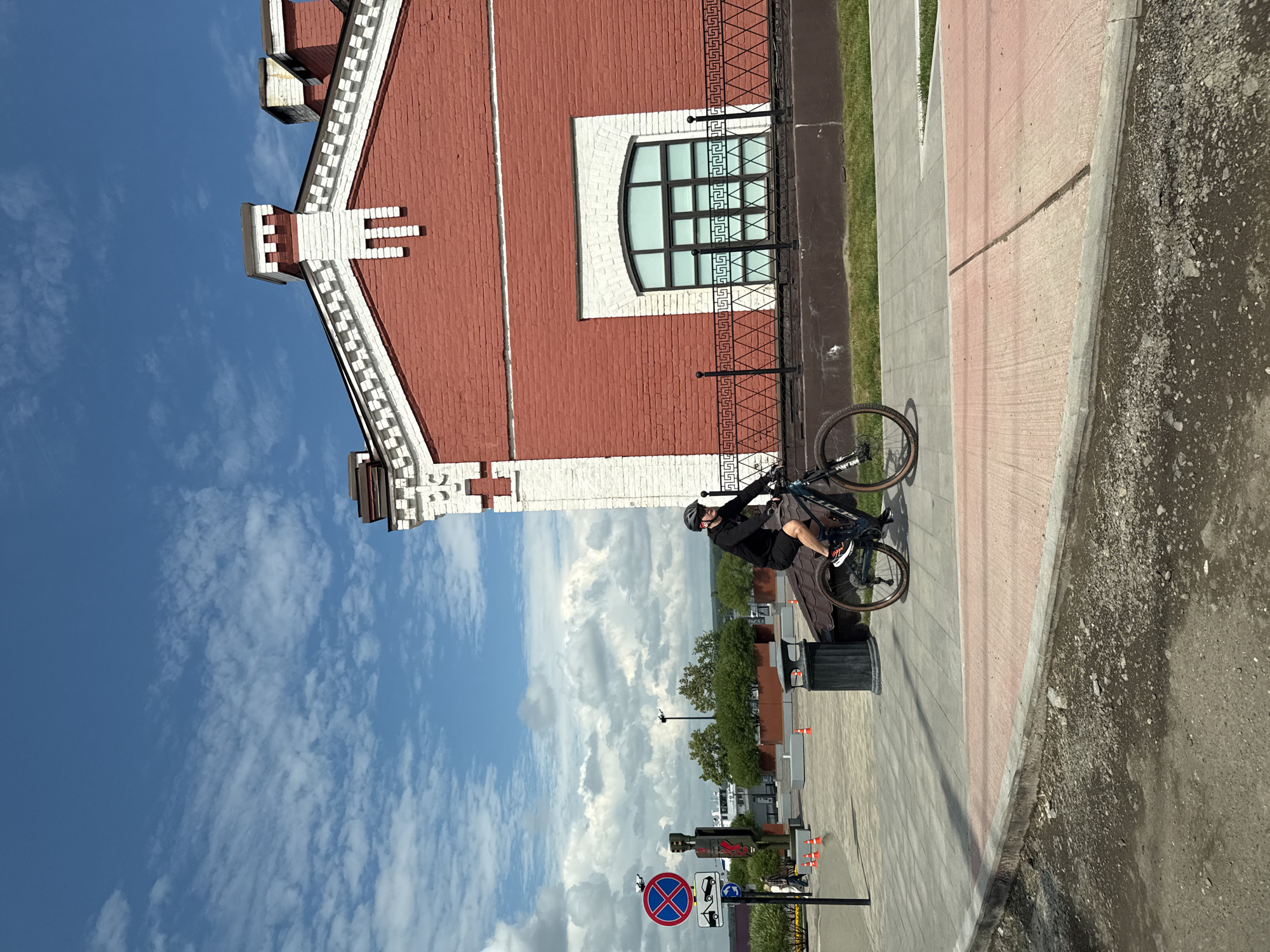 A person wearing a helmet and black athletic clothing is riding a mountain bike on a paved path. The building behind the cyclist is constructed of red brick with white decorative elements around the roofline and a large arched window. To the left of the building, there is a traffic sign indicating "No Parking" and "No Stopping." In the background, there are trees and a body of water, suggesting the path is likely along a waterfront. The sky is blue with scattered white clouds. This scene is located in the city of Perm, Russia, specifically on the embankment of the Kama River.