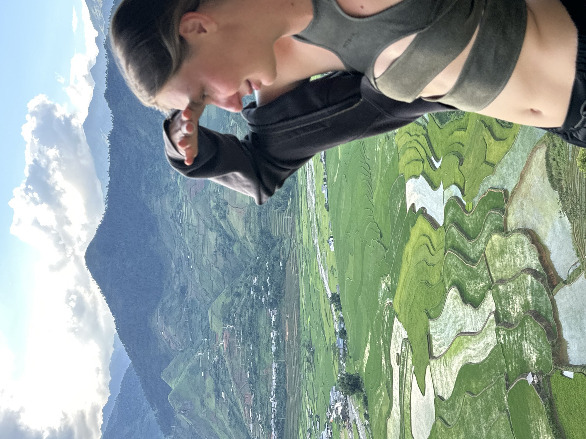 A young person, visible from the midriff up and positioned on the right side of the frame, stands on a high vantage point overlooking the Tú Lệ valley in Vietnam. The individual is facing right, with their right hand raised to their forehead, shielding their eyes as they gaze into the expansive landscape. They are wearing a dark grey open zip-up hoodie partially draped over their left shoulder, revealing a dark green, cut-out style crop top. Their midriff and navel are visible. The background presents a vast panoramic view dominated by undulating mountains, some densely covered in green foliage, under a partly cloudy sky. The valley floor is characterized by an intricate pattern of vibrant green terraced rice paddies, many filled with reflective water. A river or stream winds through the valley, bordered by scattered clusters of houses and a visible network of roads ascending the mountain slopes.
