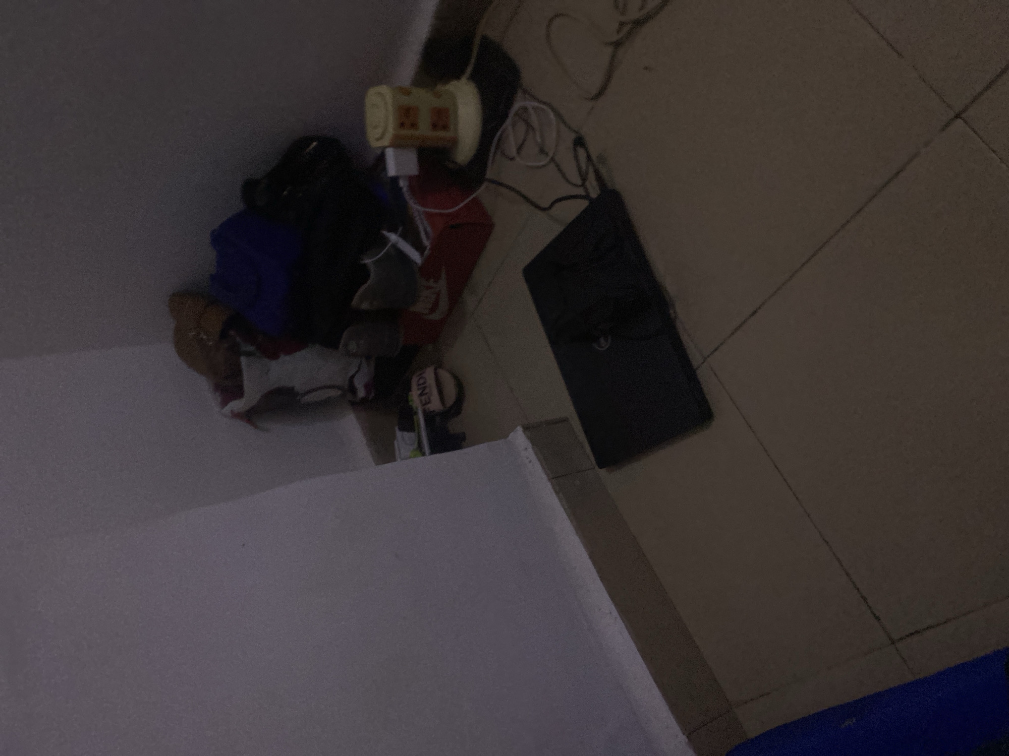 The image depicts a dimly lit corner of a room, likely a domestic setting. The floor is tiled with square, beige tiles. Against the wall, a collection of items is visible, including a red Nike shoe box, a pair of white and grey sneakers, a blue bag, and a brown bag. To the right, a cylindrical object, possibly a small lamp or fan, sits on a black mat. A laptop is placed open on the floor in the foreground, with a black computer mouse on top of it. Several white wires are tangled near the cylindrical object and the laptop. A single slipper with "FENDI" branding is visible on the floor near the shoe box. The walls appear to be painted white, with some subtle texture. There are no people present in the image. The scene suggests a casual and somewhat disorganized living space. There is no information in the image to suggest a specific location within Howayi, Nigeria; this is simply a generic interior scene.