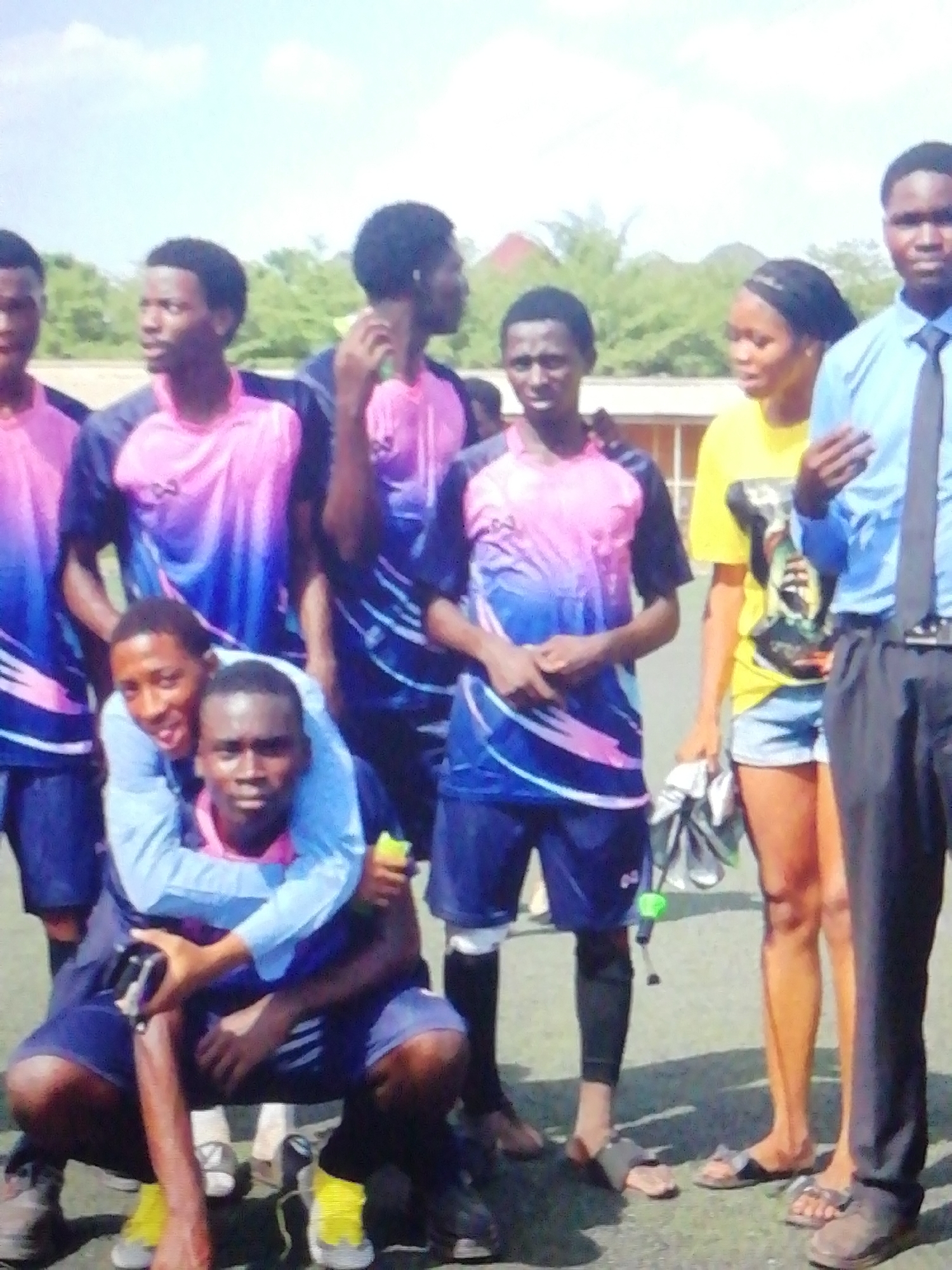 The image displays a group of ten young individuals gathered outdoors on what appears to be a grassy sports field, likely in Enugu, Nigeria. The scene suggests a sunny day, with a clear blue sky and some white clouds overhead, indicating a midday or afternoon setting.
The subjects include eight young men and two young women. Most of the men are wearing matching short-sleeved soccer jerseys that feature a distinctive pink and blue gradient pattern, with blue shorts. On the chest of at least one jersey, the word "TCO" is visible. One man wears a light blue long-sleeved shirt, piggybacking another who is crouching, also in blue shorts. Another man stands to the right, dressed in a light blue button-up shirt, a black tie, and black trousers, appearing more formally dressed. The two young women are on the right side of the frame; one wears a bright yellow short-sleeved t-shirt with a graphic design and denim shorts, while the other's attire is less clear.
The group appears to be posing for a photograph, with various expressions ranging from neutral to slight smiles. The man in the light blue long-sleeved shirt has a more pronounced smile. Some individuals are barefoot, others wear sandals or sneakers. In the background, green foliage from trees and some distant building structures are visible. The ground is a green turf or well-maintained grass. The overall atmosphere suggests a casual gathering, possibly after a sports event or practice.