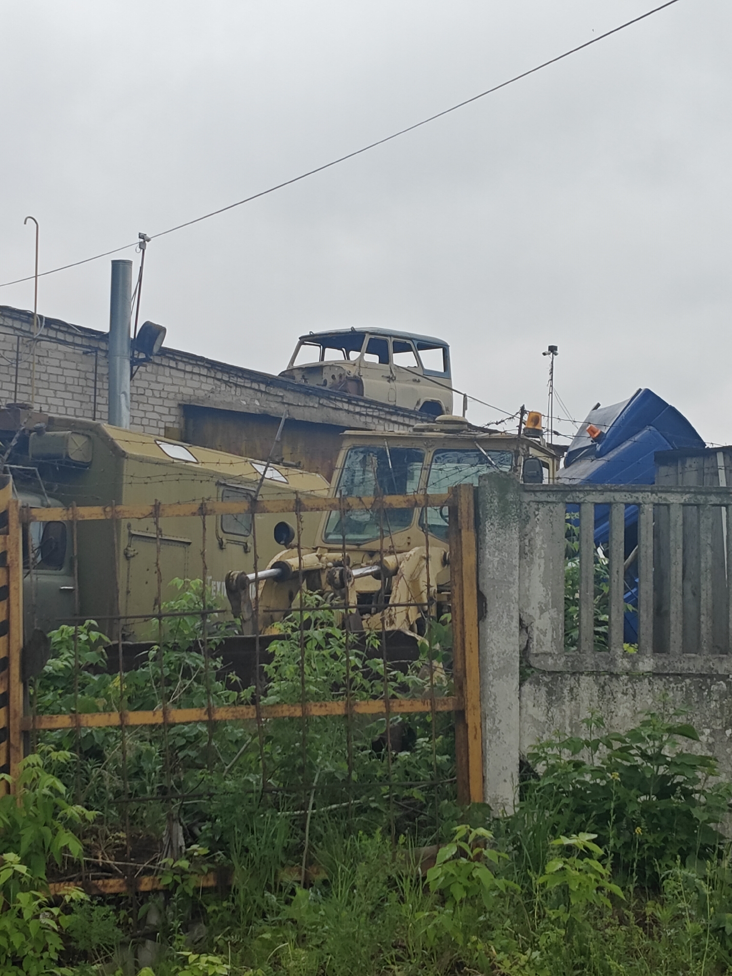 The image depicts an outdoor scene, likely a scrapyard or storage area in Chaikovsky, Russia, given the vehicles present. Several derelict vehicles are visible. One vehicle appears to be an armored personnel carrier or similar military vehicle, while another seems to be the stripped chassis of a truck or other larger vehicle. A fence made of yellow metal and concrete forms a barrier in the foreground, partially obscured by overgrown vegetation. A brick building is also visible in the background. The overall scene conveys a sense of neglect and disuse. The sky is overcast, which suggests it is daytime.
