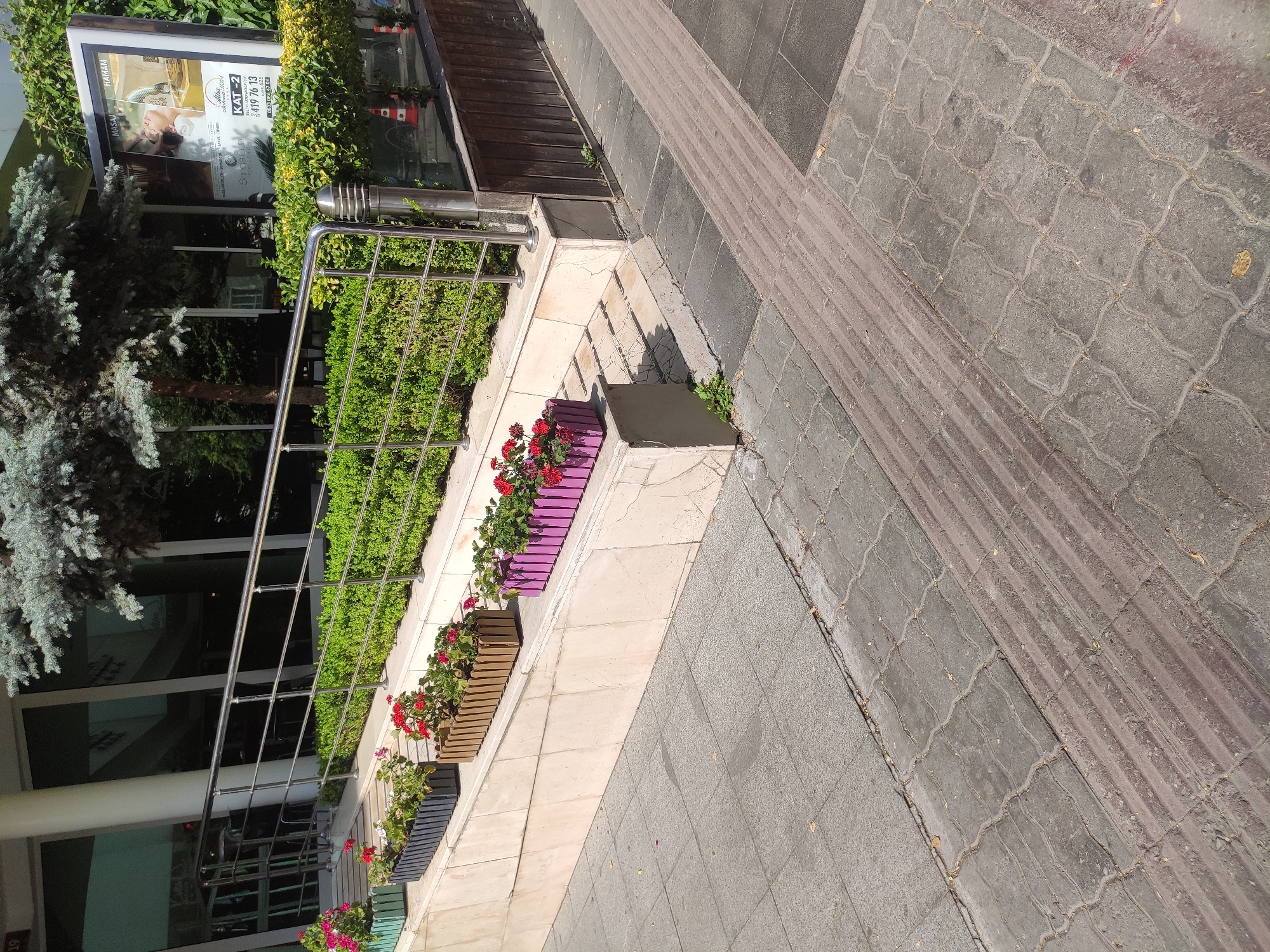 The image depicts an outdoor scene with a building entrance and sidewalk. A metal railing guides pedestrians up a ramp towards the building. Planters filled with red flowers are strategically placed along the ramp and on a raised concrete area. The sidewalk is paved with interlocking concrete blocks, and a textured section, likely for visually impaired individuals, runs diagonally across the pavement. In the background, a large billboard advertises "MASAJ HAMAM," suggesting a spa or wellness center. The environment appears to be a city street in Ankara, Turkey, given the context of the billboard's advertising language and the architectural style. There are no people visible in the image. The scene is well-lit by daylight, casting shadows on the pavement.