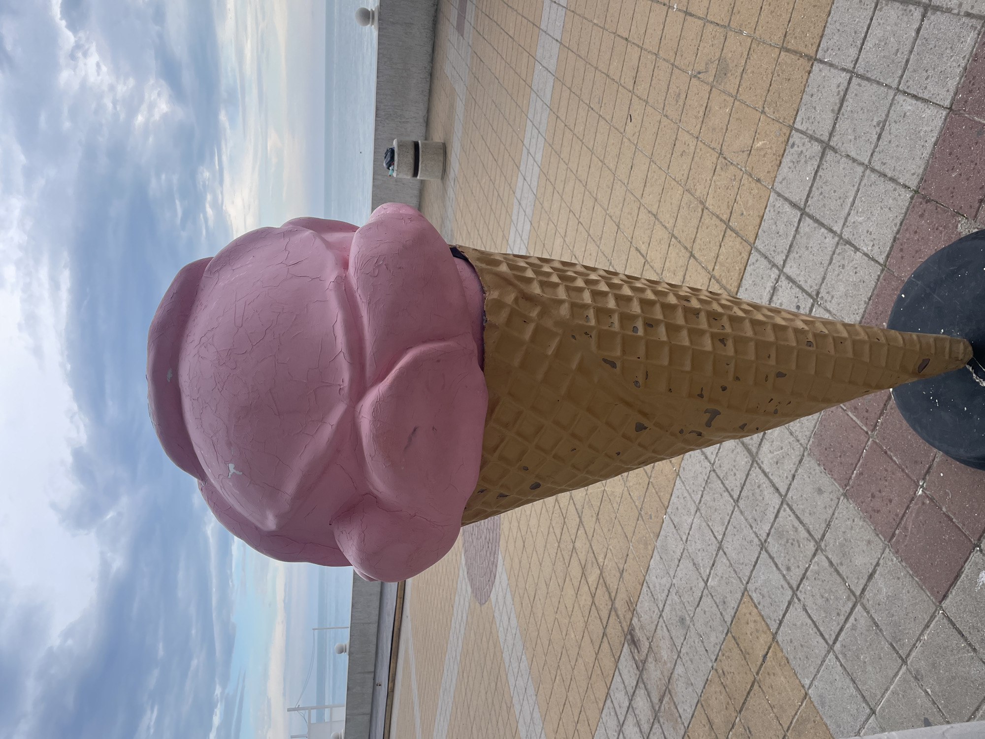 A large, pink, sculpted ice cream cone sits on a paved walkway with a view of the sea and cloudy sky. The walkway is made of beige, gray, and red square tiles. A low concrete barrier and white fence border the walkway. A metal trash can is visible in the background. The ice cream cone has a cracked paint finish on the pink scoop and a waffle pattern on the cone. A black circular base is beneath the cone. This is in the city of Olginka, Russia.