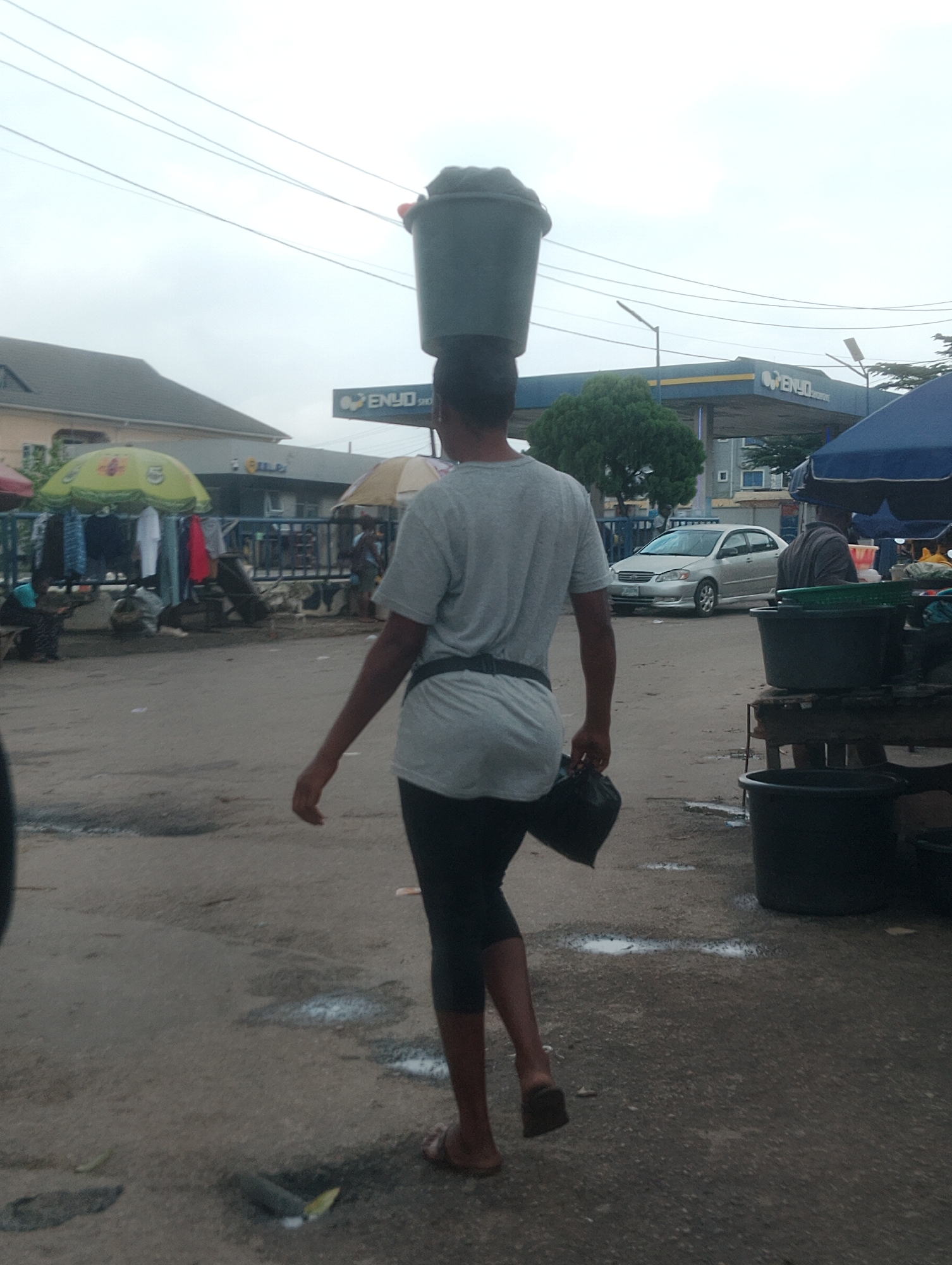 A person walks across a somewhat wet, dusty ground in Iju, Nigeria. They are carrying a large, dark-colored bucket balanced on their head. The person is wearing a light grey t-shirt, black leggings or tight pants, and sandals. A black bag is held in their right hand. The background features a gas station with the sign "ENYO" clearly visible. There are also market stalls with clothes displayed under umbrellas, a silver car, and several large black plastic containers. The sky is overcast, suggesting it might be a cloudy day or nearing evening. The overall scene appears to be a busy, everyday street market or roadside commerce area.