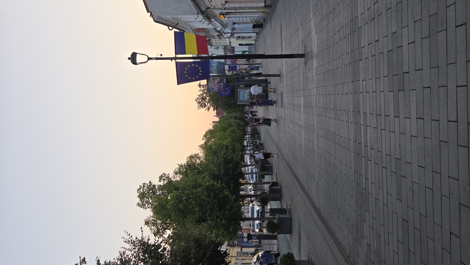 The image depicts a street scene in Cluj-Napoca, Romania, likely in the city center during the late afternoon or early evening. The perspective is from the pedestrian walkway looking down a street lined with trees and buildings.
On the left side of the street, a row of mature trees in square planters creates a verdant canopy. Cars are parked along the curb and traffic is moving on the road. Further down, a blue car is visible.
The pedestrian walkway is paved with rectangular gray bricks laid in a herringbone pattern. Several people are walking along the walkway. On the right side of the walkway, a tall black lamppost is prominently featured, flying the European Union flag and the Romanian flag. Behind the lamppost, buildings with white facades and arched windows line the street. Several advertisements and posters are displayed on poles along the walkway. The sky is a pale orange and blue gradient, indicating the sunset.