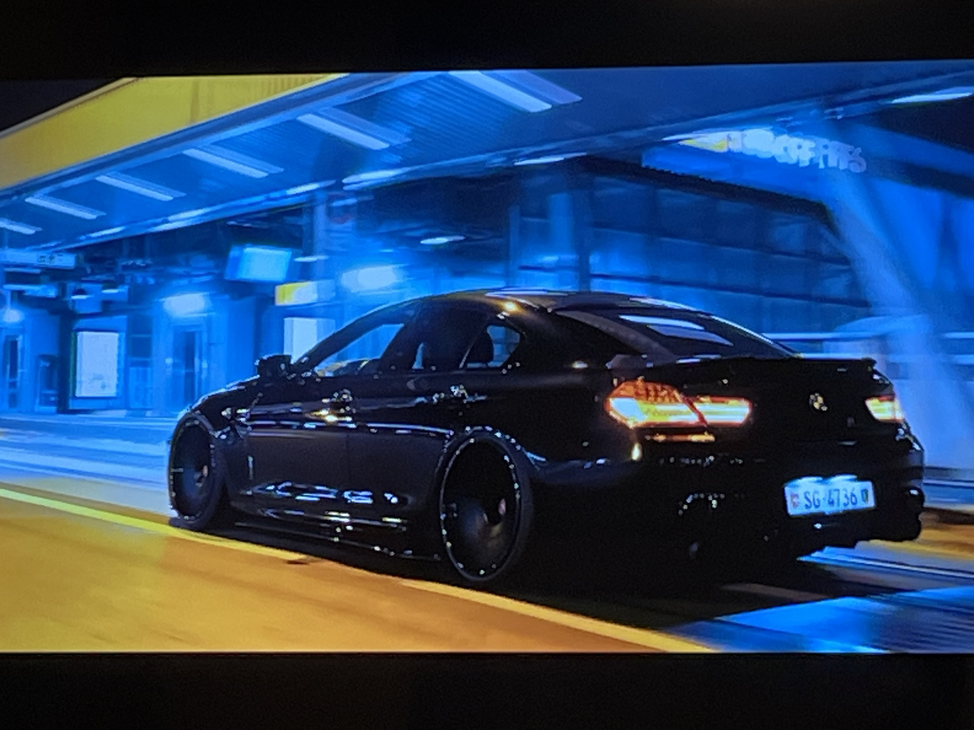 The image shows the rear passenger-side view of a black sedan driving on a wet, yellow-lined road at night. The car has custom black rims and a license plate that reads "SG 4736". The background is blurred due to motion, but appears to be an urban environment with blue-lit structures that resemble a train station or covered walkway. There are no people visible in the image. The car's taillights are illuminated. The location is identified as Kharkiv, Ukraine.