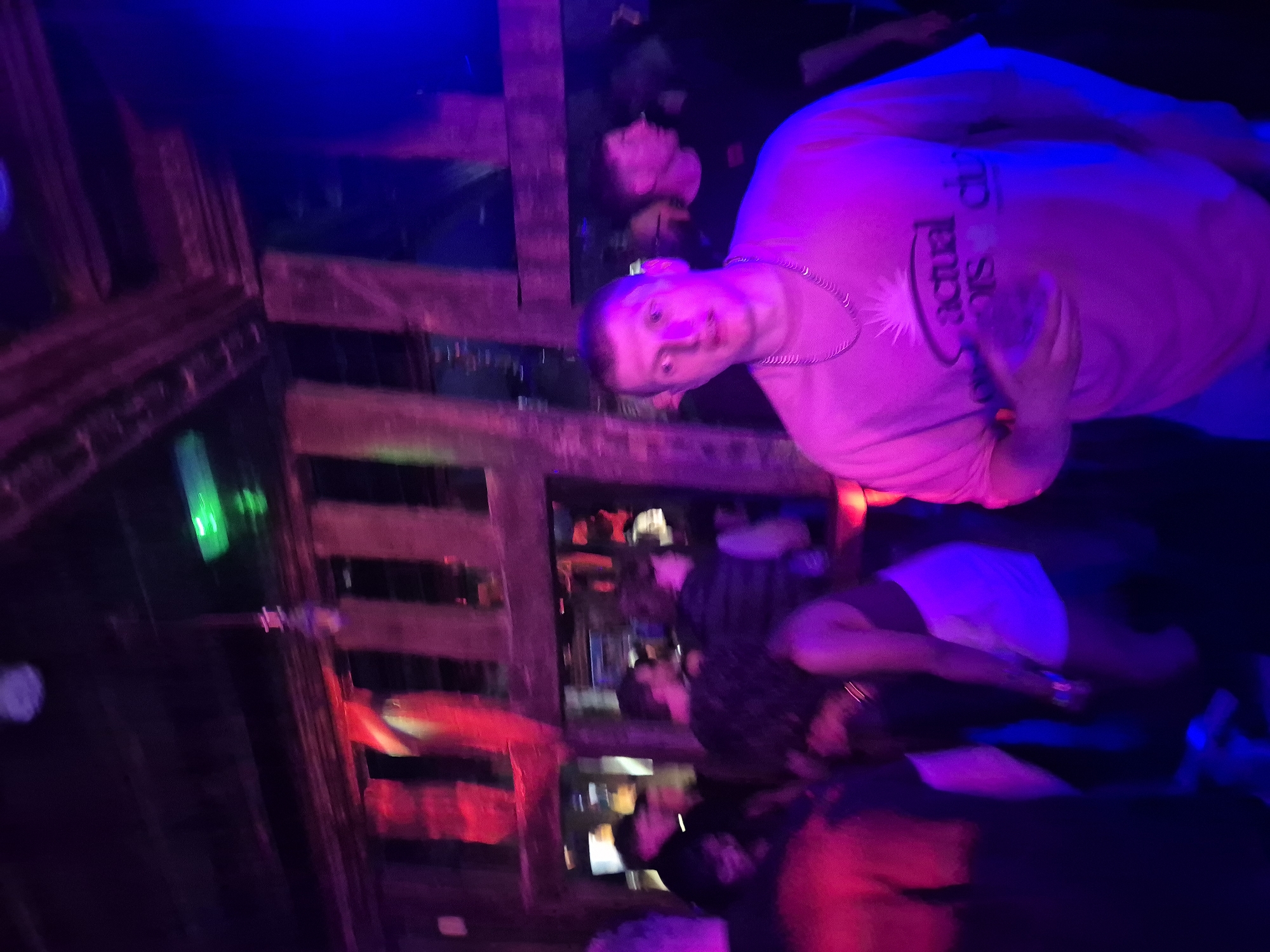 The scene appears to be inside a dimly lit bar or nightclub, possibly in West Suffolk, United Kingdom, based on implied contextual information (no specific location details provided in the image). The primary subject is a fair-skinned man with a short haircut, wearing a pink t-shirt and a silver chain necklace. He is holding a clear glass, likely containing a drink. The text on his t-shirt appears to read "Actuarial club". The man is looking towards the camera.
Behind him, there are several other people, and a possible bar area with shelves stocked with bottles. The lighting is a mix of blue, red, and green, creating a nightclub atmosphere. The perspective of the photo is somewhat skewed and blurry. There is visible wood decor on the walls.