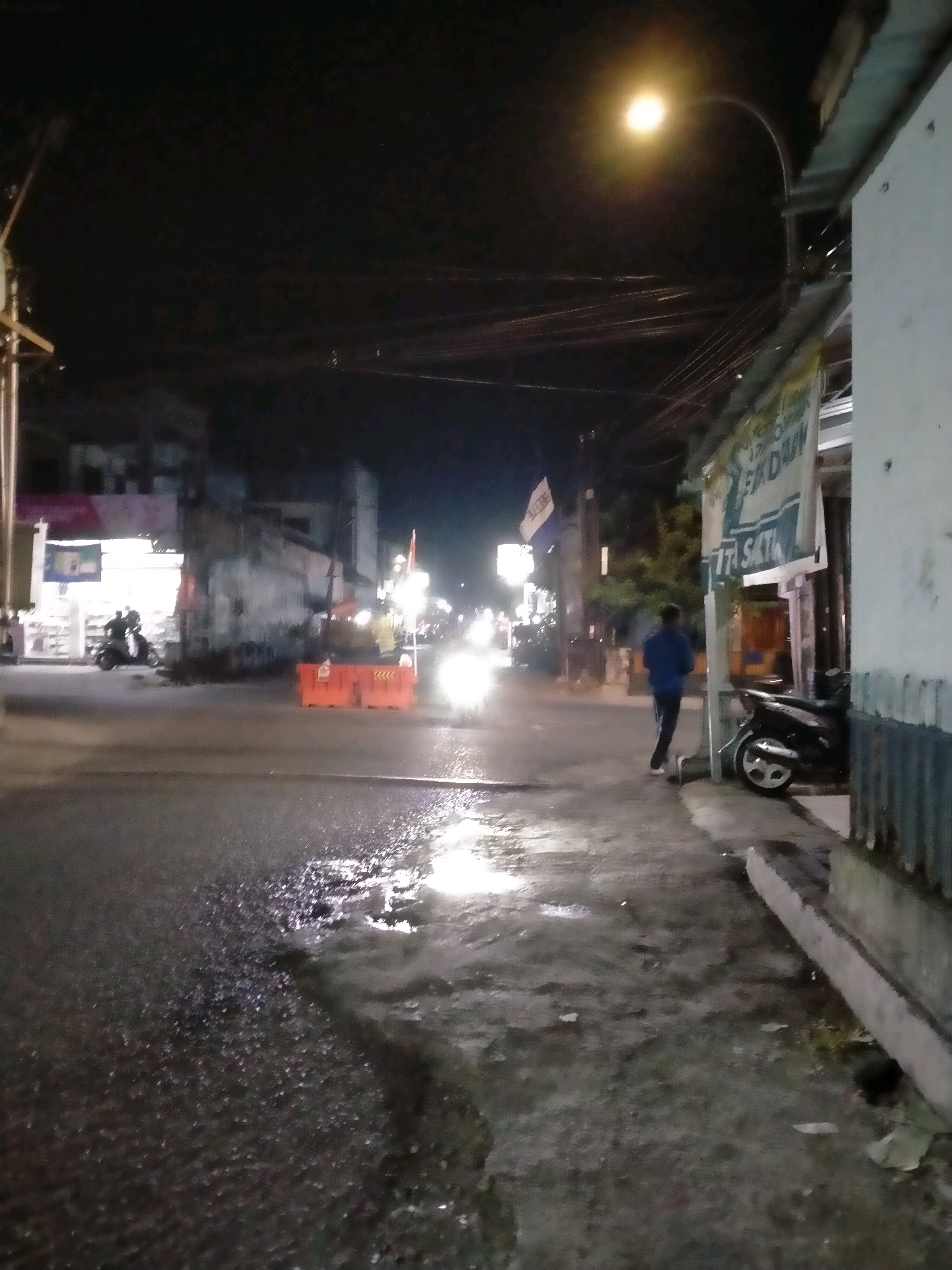 A night-time, street-level view captures a scene in the city of Kasihan, Indonesia, under dim artificial lighting. The asphalt road appears wet and reflects the light sources. In the foreground, the wet surface is textured with small, reflective particles. To the right of the frame, a narrow sidewalk runs alongside a building with a corrugated metal exterior. A motorcycle is parked on the sidewalk. A person, wearing a blue jacket and dark pants, stands on the sidewalk facing away from the camera, towards the street. Further down the street, to the left, a store or business with illuminated windows is visible, with a person on a scooter in front of it. Orange traffic barriers are positioned in the middle of the road, suggesting ongoing construction or a closed-off area. The sky is dark, with visible overhead wires and a streetlamp casting a bright glow. The scene is characterized by a general blurriness, indicative of low light conditions and potentially camera movement.