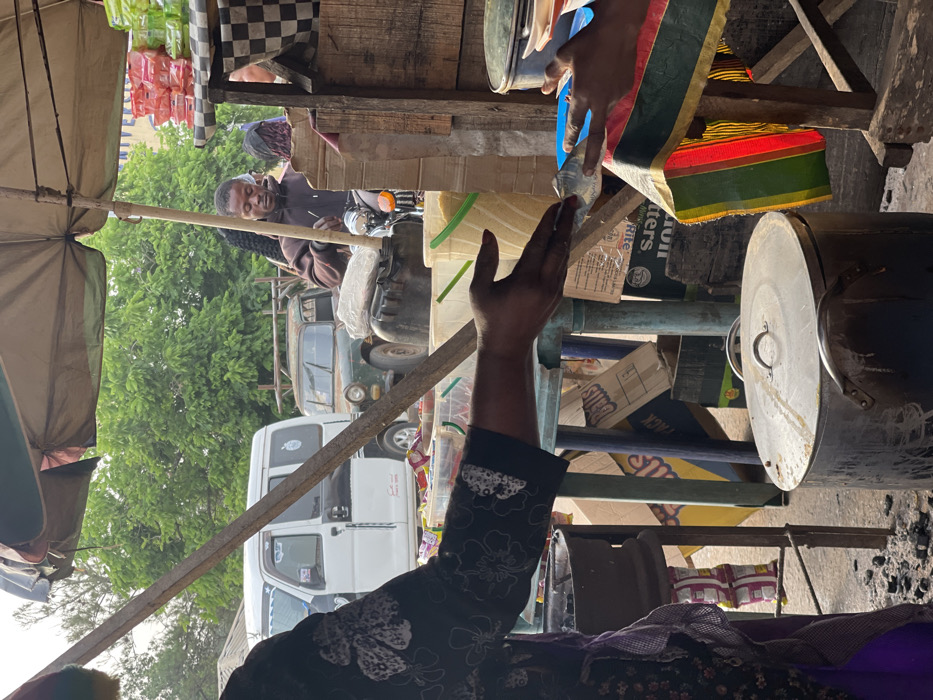 A market scene features a man on a motorcycle, smiling under a checkerboard awning. A white van and an older, green vehicle are visible behind him. In the foreground, a person's arm is raised in a greeting or gesture. Stacks of boxes, cooking pots, and various goods fill the frame, suggesting a bustling commercial environment. Colors, patterns, and objects are layered, creating a dense and vibrant composition.