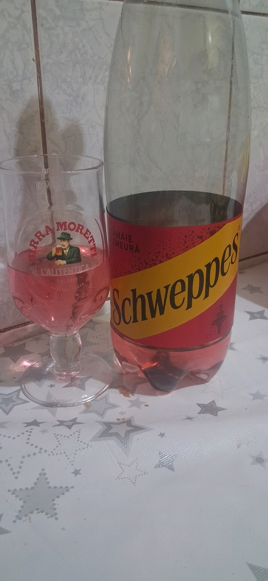 This is a close-up, eye-level, indoor shot of a glass and a plastic bottle, likely on a table in Satu Mare, Romania. The glass, to the left, is partially filled with a pink liquid and has "BIRRA MORETTI" printed on it, along with a picture of a man in a green hat holding a beer. Next to the glass, to the right, is a large plastic bottle of Schweppes. The bottle has a red label with a yellow diagonal stripe and the Schweppes logo. The background appears to be a tiled wall. The table surface is white with a repeating pattern of grey stars. There is no visible activity or interaction occurring.
