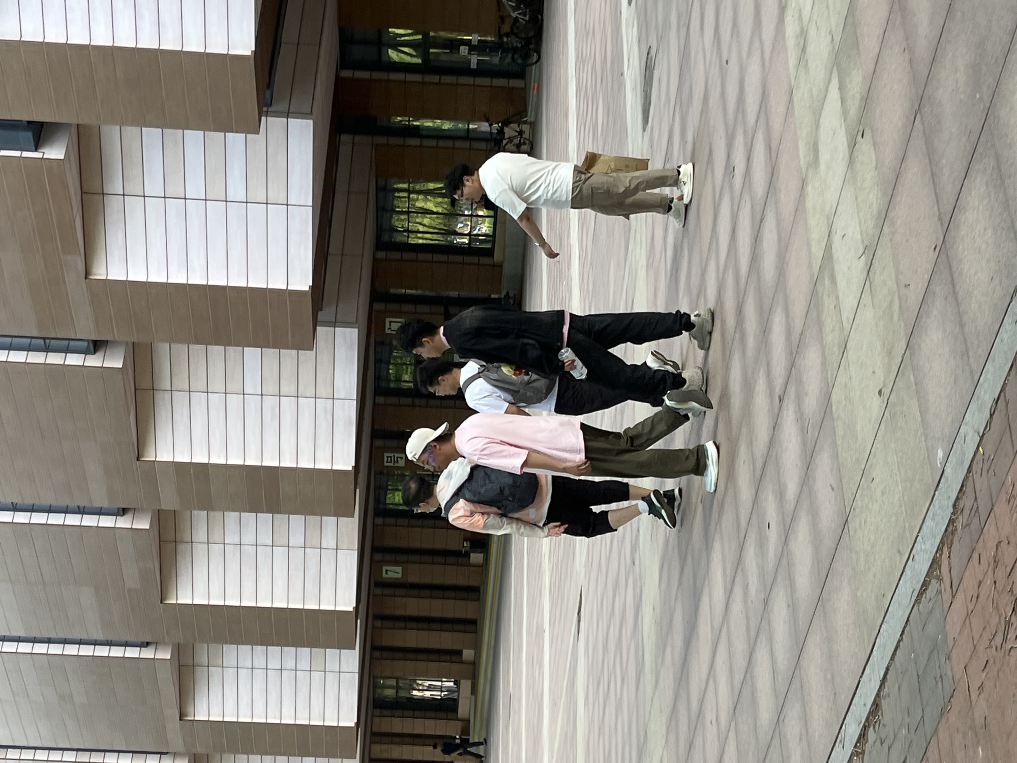The image shows a group of four young people walking on a tiled plaza in Changchun City, China. They are walking away from the camera and away from a building with a layered rectangular facade. A person is walking off to the left of the building and another person is to the right of the four young people. The plaza is tiled in a grid pattern. The overall setting is an urban environment, likely near a university or public building, given the architectural style.