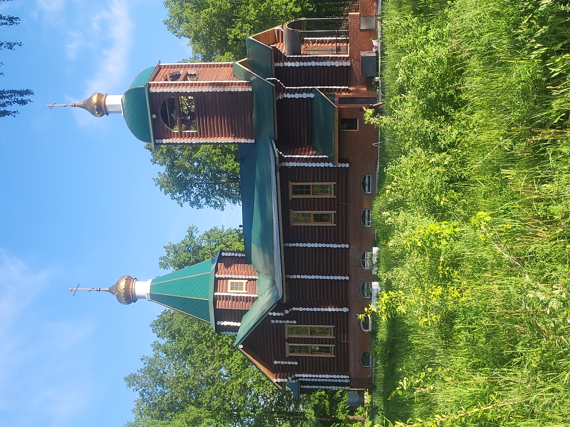 The image depicts a wooden Russian Orthodox church with green roofs and golden domes, set amongst lush green trees and foliage. The church features detailed wood carvings and white trim, with a prominent bell tower topped with a dome and cross. The building's architecture appears traditional, showcasing intricate craftsmanship. The location is identified as Chaikovsky, Russia, based on visual features characteristic of Russian Orthodox church architecture. The foreground is filled with tall grass and dense vegetation, suggesting a natural setting or park. The sky is partially visible, revealing a clear blue expanse with scattered clouds.
