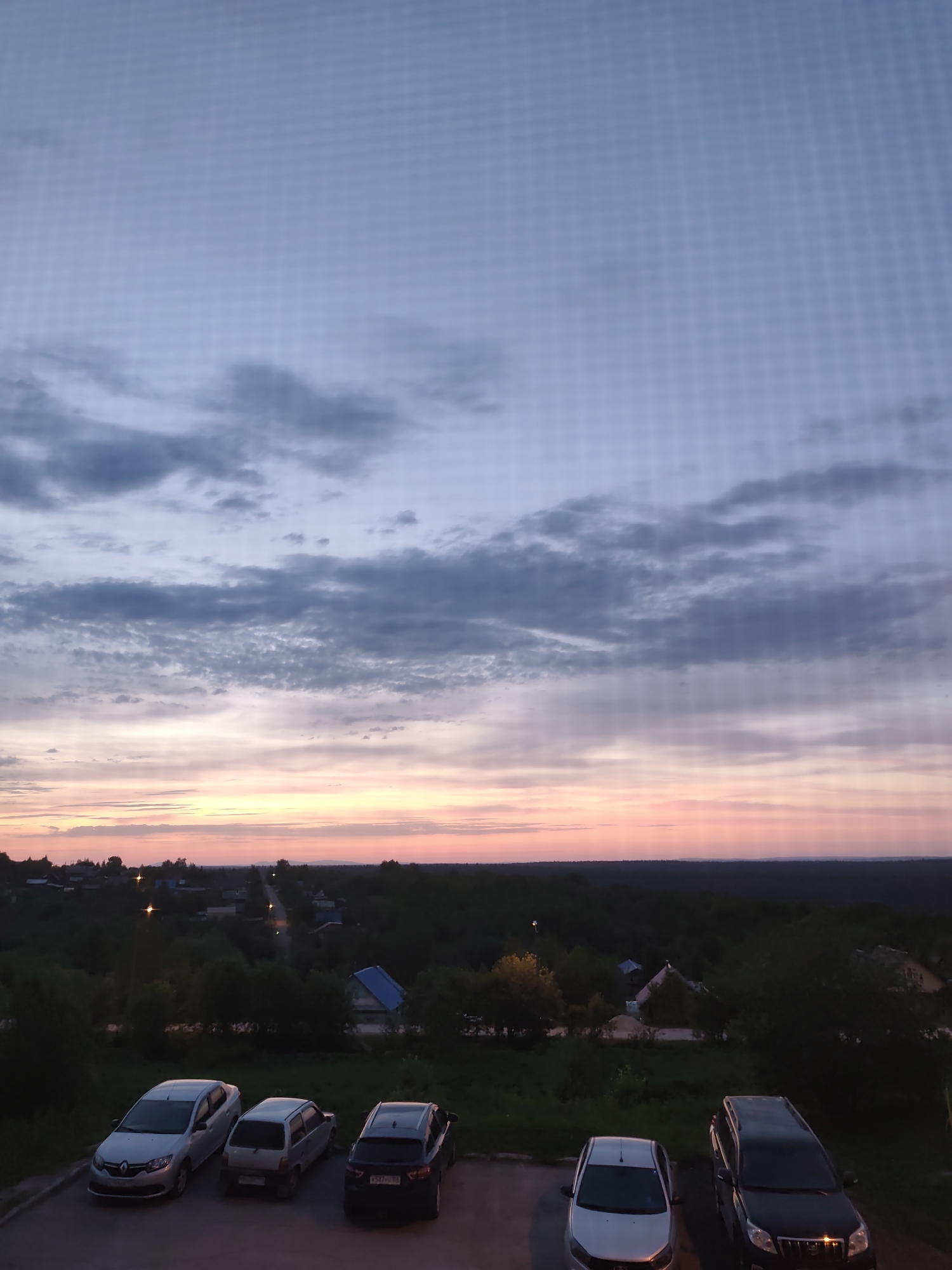 The image shows a view from an elevated position overlooking a landscape at dusk. The sky transitions from blue-grey to pink and orange near the horizon, indicating a sunset or sunrise. There are scattered clouds in the sky. Below, a dark, dense collection of trees obscures much of the ground, but some rooftops and streetlights are visible, suggesting a residential area or small town. In the foreground, there is a parking area with five cars: a silver Renault, a silver small car, a black SUV, a silver sedan, and a black SUV.
While the prompt mentions Gremyachinsk, Russia, there's nothing visually in the image to confirm this location definitively.