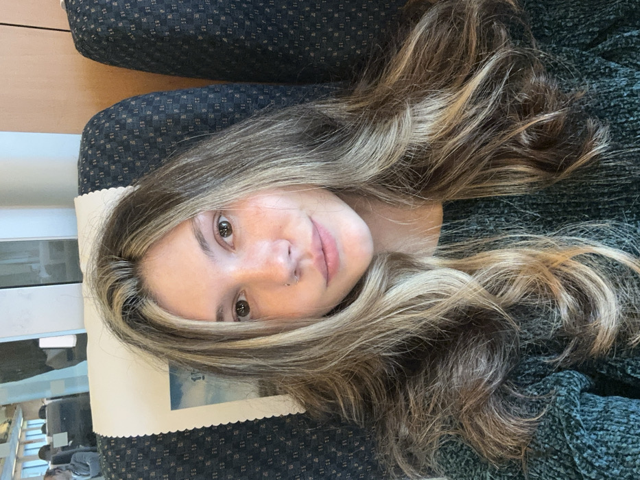 A female subject is centrally positioned in a close-up, frontal view within a passenger train car. She has long, wavy, multi-toned hair, predominantly brown with prominent blonde highlights, and dark brown eyes. A small stud piercing is visible in her left nostril. She wears a dark green, textured knit sweater and maintains a neutral expression with closed lips, looking directly at the viewer. She is seated in a dark blue patterned train seat, which features a white, scalloped-edge headrest cover behind her left shoulder. A partial blue and white image is visible on the headrest cover. In the background, faint reflections and partial views of other passengers and windows are discernable, indicating the interior of a moving train. The scene is located within Tonglu County, China.
