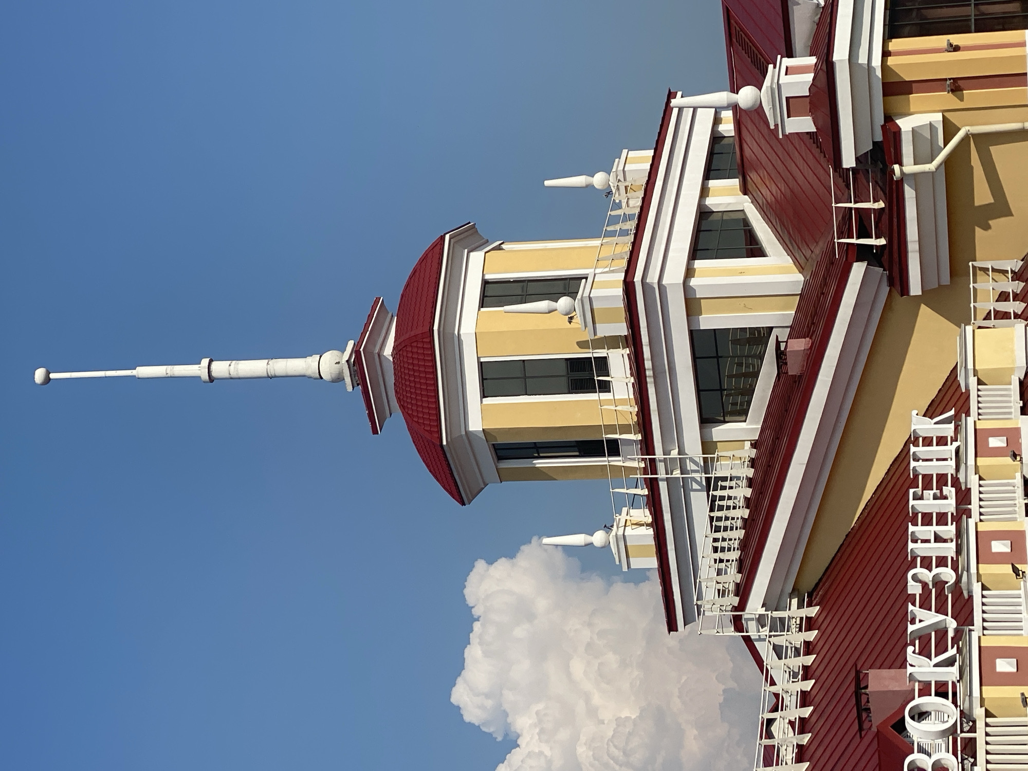 This is a high-angle, close-up photograph of the Novokuznetsk railway station building in Novokuznetsk, Russia, on a clear day with a large white cloud in the sky. The building features a distinctive red roof and yellow facade, with white decorative elements. A tall, slender white spire tops the central section of the building. White metal railings and a ladder are visible on the roof. Large white letters spelling out "ВОКЗАЛ" (Vokzal, meaning "station" in Russian) are mounted on the lower section of the building. The sky is a vibrant blue. There are no people visible in the image.