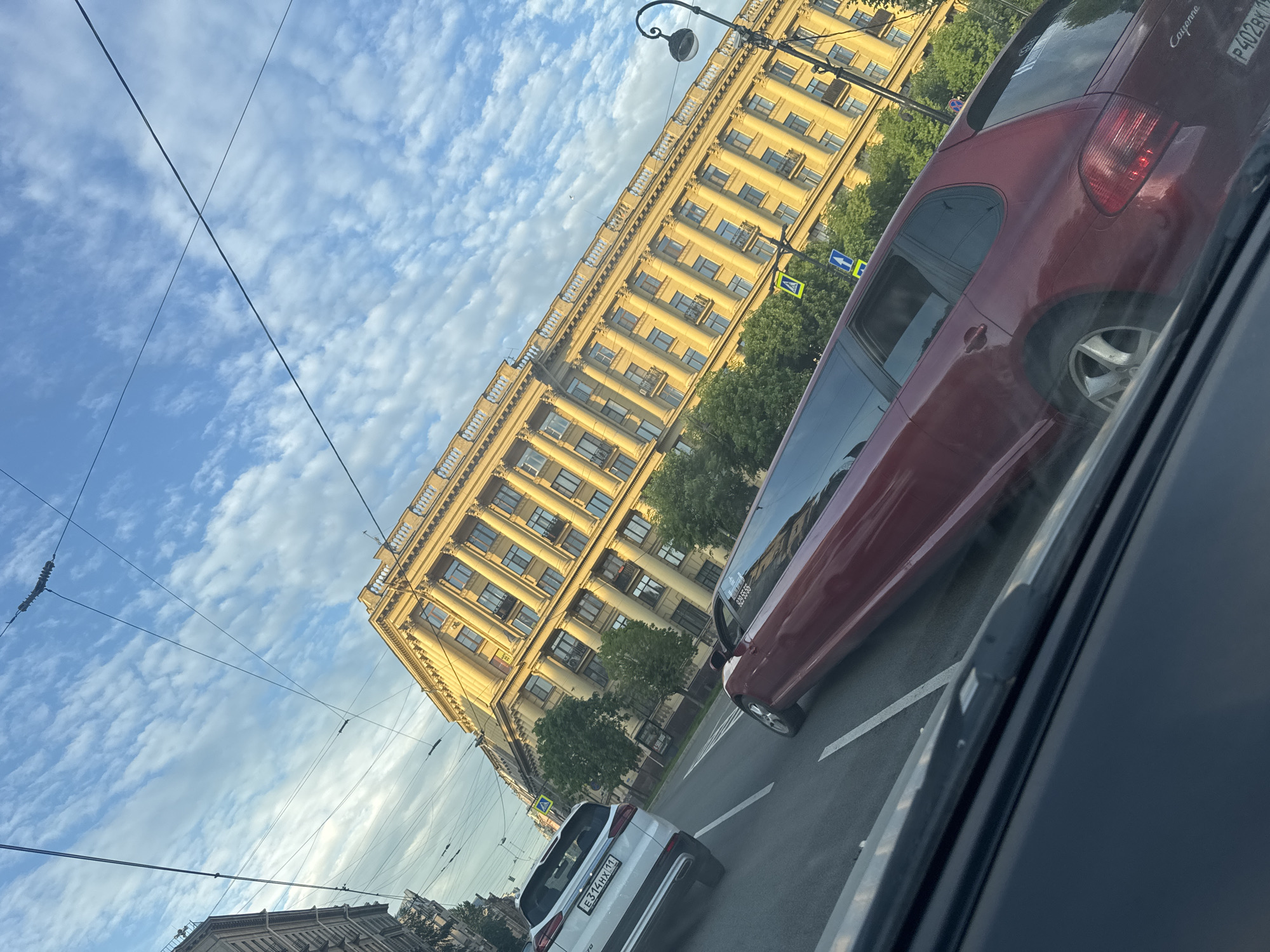 The image captures a tilted, sunlit street scene in Saint Petersburg, Russia, viewed from inside a vehicle. A clear blue sky with scattered white cumulus clouds dominates the upper left, crossed by multiple overhead wires, possibly for trolleybuses or trams.
On the right, a grand, multi-storied building with classical architecture stands prominently. It's painted in a warm yellow, featuring numerous windows, decorative columns, and cornices. Green trees line the sidewalk in front of the building, and a traditional black street lamp extends from its facade.
In the foreground, a dark red Porsche Cayenne SUV is seen from its rear and side, with the model name "Cayenne" visible above its right taillight. A Russian license plate, partially obscured, shows "P402BK". Further down the street, a white car is visible, bearing a Russian license plate "E314HX 111". Both cars are on a paved road with white markings, suggesting a city street.
Visible street signs include two blue rectangular signs with white arrows (indicating directions) and a yellow diamond-shaped sign, likely for a pedestrian crossing. The bright natural light indicates it is daytime with clear weather. The overall scene depicts a typical urban environment in Saint Petersburg, combining historic architecture with modern street traffic.