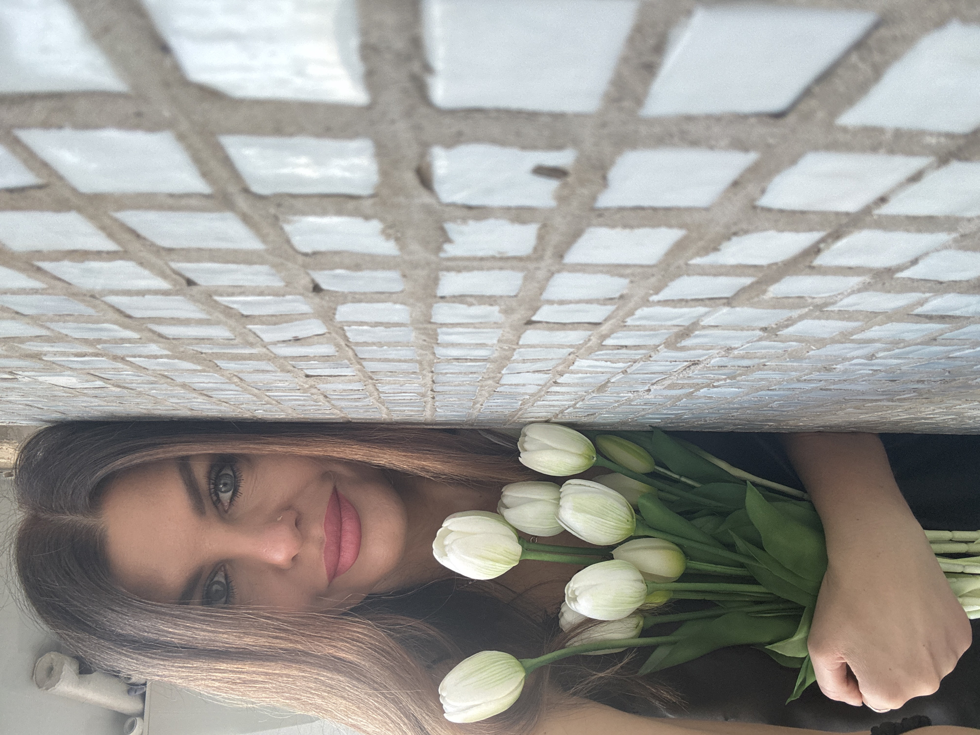 A woman with light brown hair, fair skin, and pink lipstick is lying down, holding a bouquet of white tulips. Her head is positioned near a wall tiled with small, square, off-white tiles. The background is blurred. There is no explicit information indicating that the location is Chaikovsky, Russia.