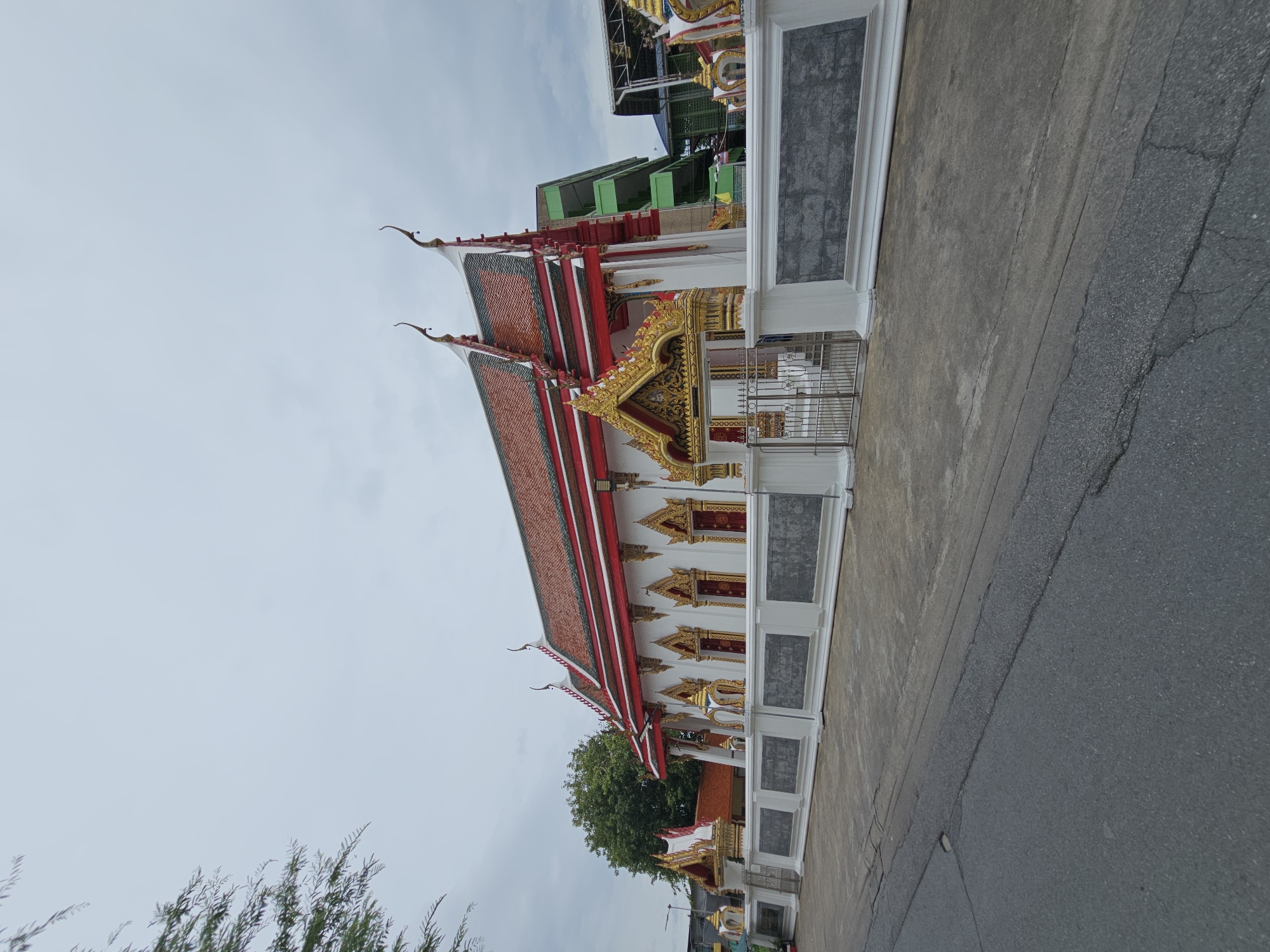 An exterior, eye-level, full shot shows a white Buddhist temple with a red, multi-tiered roof adorned with golden, ornate decorations. The temple is bordered by a white stone wall with intricate grey carvings, and a silver metal gate is visible in the center. Behind the temple, a tall, green, multi-story building can be seen, suggesting an urban environment. The sky is overcast and cloudy, and a paved road runs in front of the temple. A tree with green foliage is on the left side of the frame.