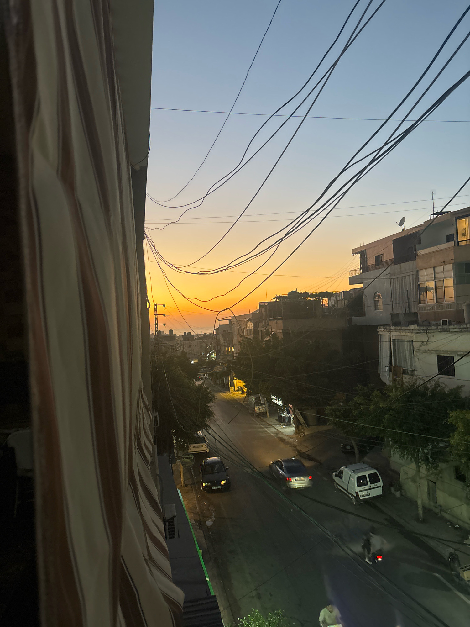A street scene in Al Bazuriya, Lebanon, captures the warm hues of sunset casting an orange glow across the sky. A building with striped awnings is visible in the foreground on the left. Several cars are present on the asphalt road, including a black sedan, a silver sedan, and a white van. Overhead, a tangle of power lines crisscrosses the sky. Trees and other buildings line the street, some with illuminated windows. The overall atmosphere suggests an urban environment at dusk.
