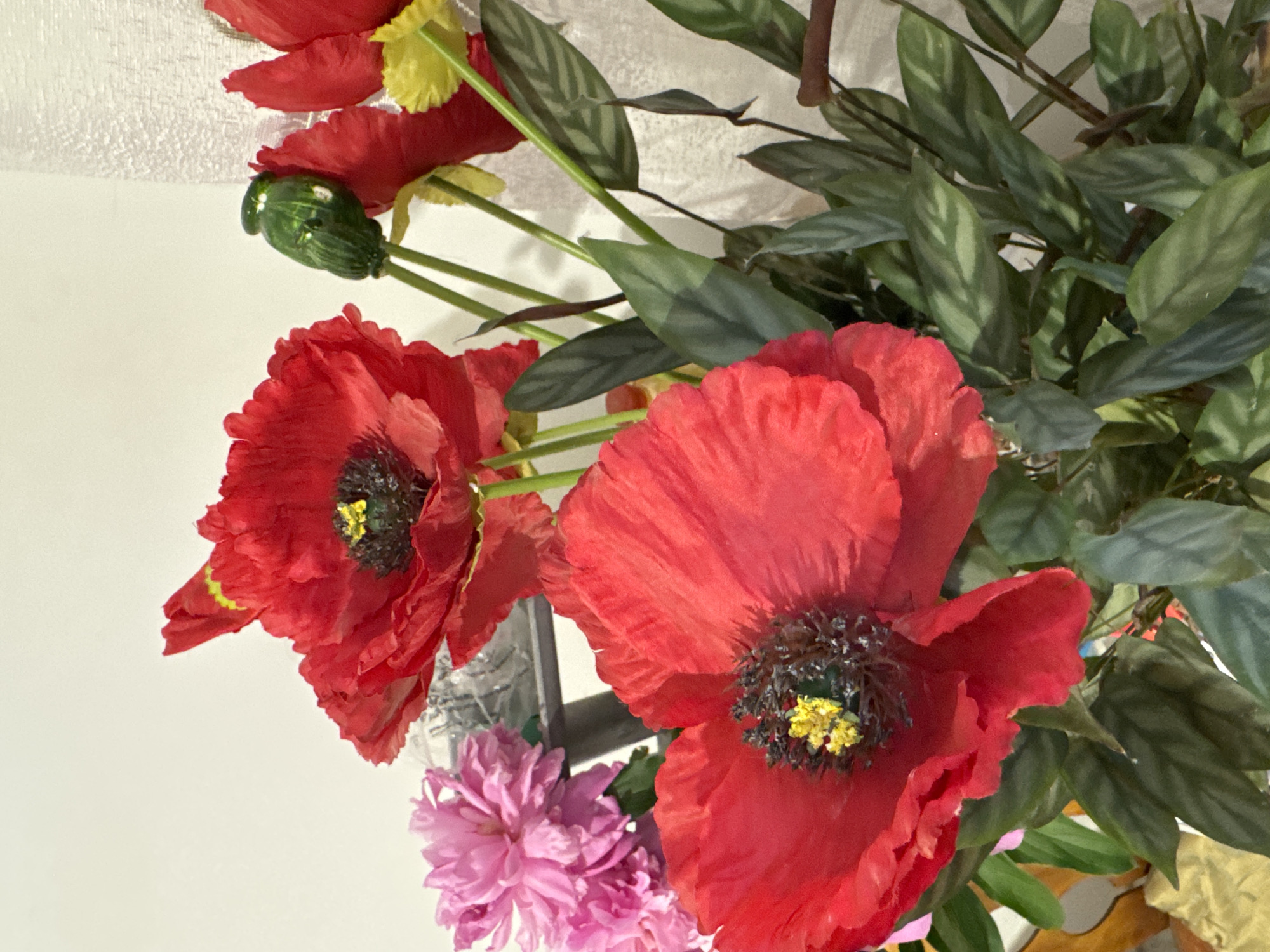 The image shows a close-up of a floral arrangement featuring several red poppies, some pink flowers, and green foliage. One of the poppies in the foreground is particularly prominent, with its vibrant red petals and dark center with yellow stamen. Other red poppies are visible in the background, along with a green poppy bud and a yellow flower. The pink flowers, possibly peonies, are clustered to the left of the frame. The leaves are a deep green with lighter green veining. The background is a plain white wall, with a textured surface on the right side. There is no indication of the city of Kazan, Russia, in the image.