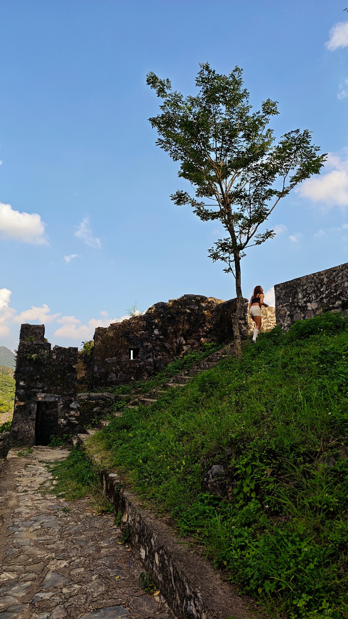 A woman walks up stone steps alongside a weathered stone wall at the Citadel of the Hồ Dynasty in Đường Thượng Commune, Vietnam. The wall, partially overgrown with grass, ascends a grassy slope leading to a tree. A crumbling stone structure with an arched opening is visible to the left. The sky is blue with scattered clouds. A paved path leads towards the stone structure.