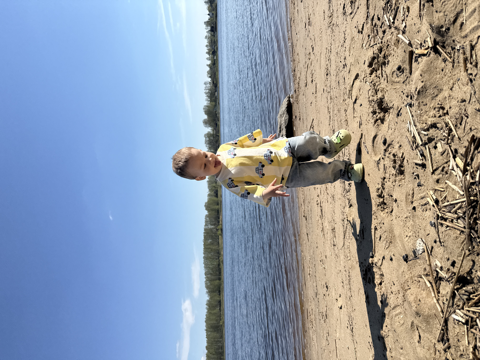 A young child is standing on a sandy beach next to a body of water. The child is wearing a yellow shirt with a car print, blue jeans, and green shoes. The water is calm with trees visible on the opposite bank. The sky is clear and blue. The sand is littered with debris. The child is holding their hands out. This image may be taken in Rusanovo, Russia, given the prompt's reference.